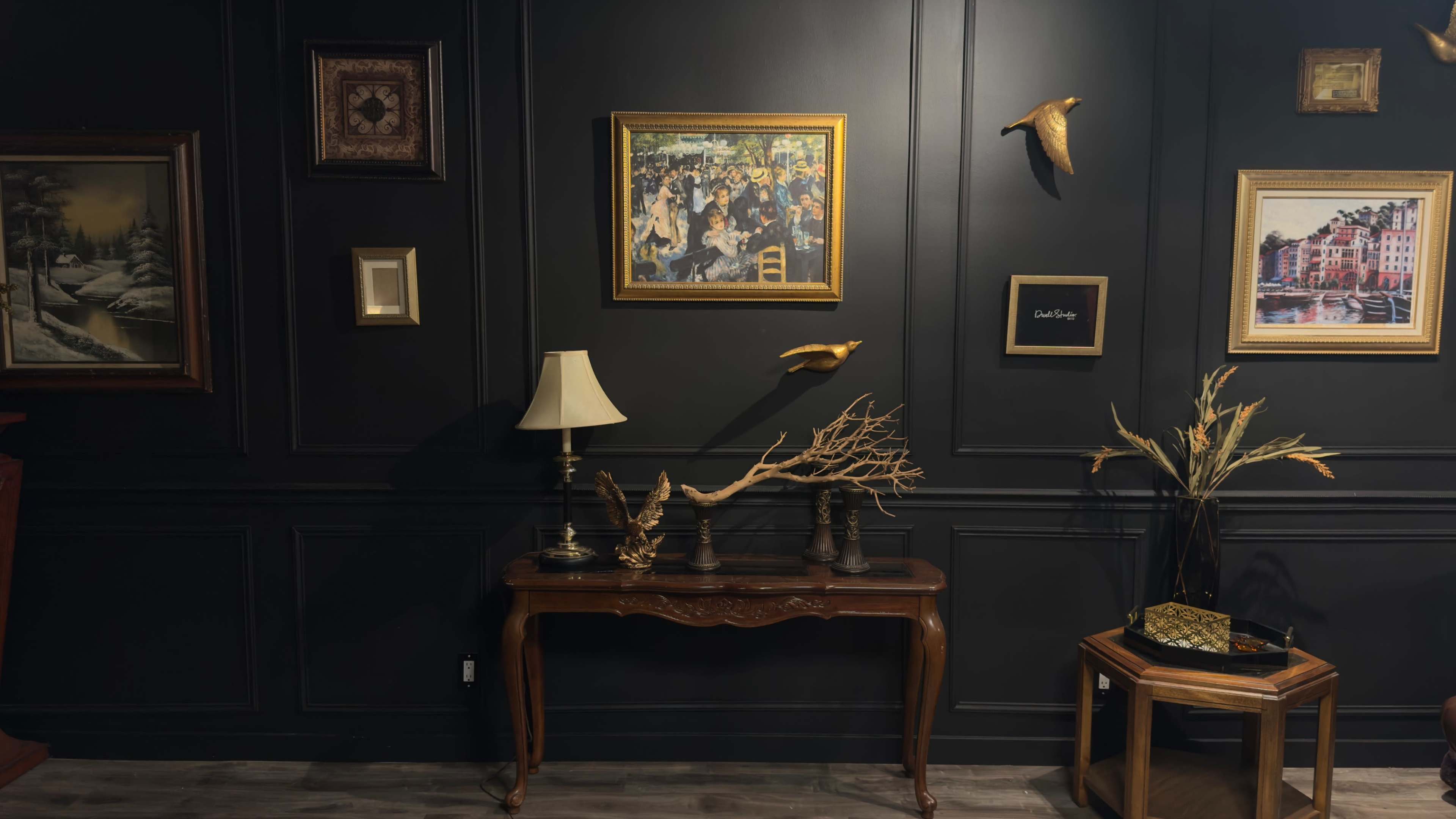 The scene features a dark-hued room with a collection of framed artwork on the walls, a decorative table with various objects, and a lamp illuminating the space.