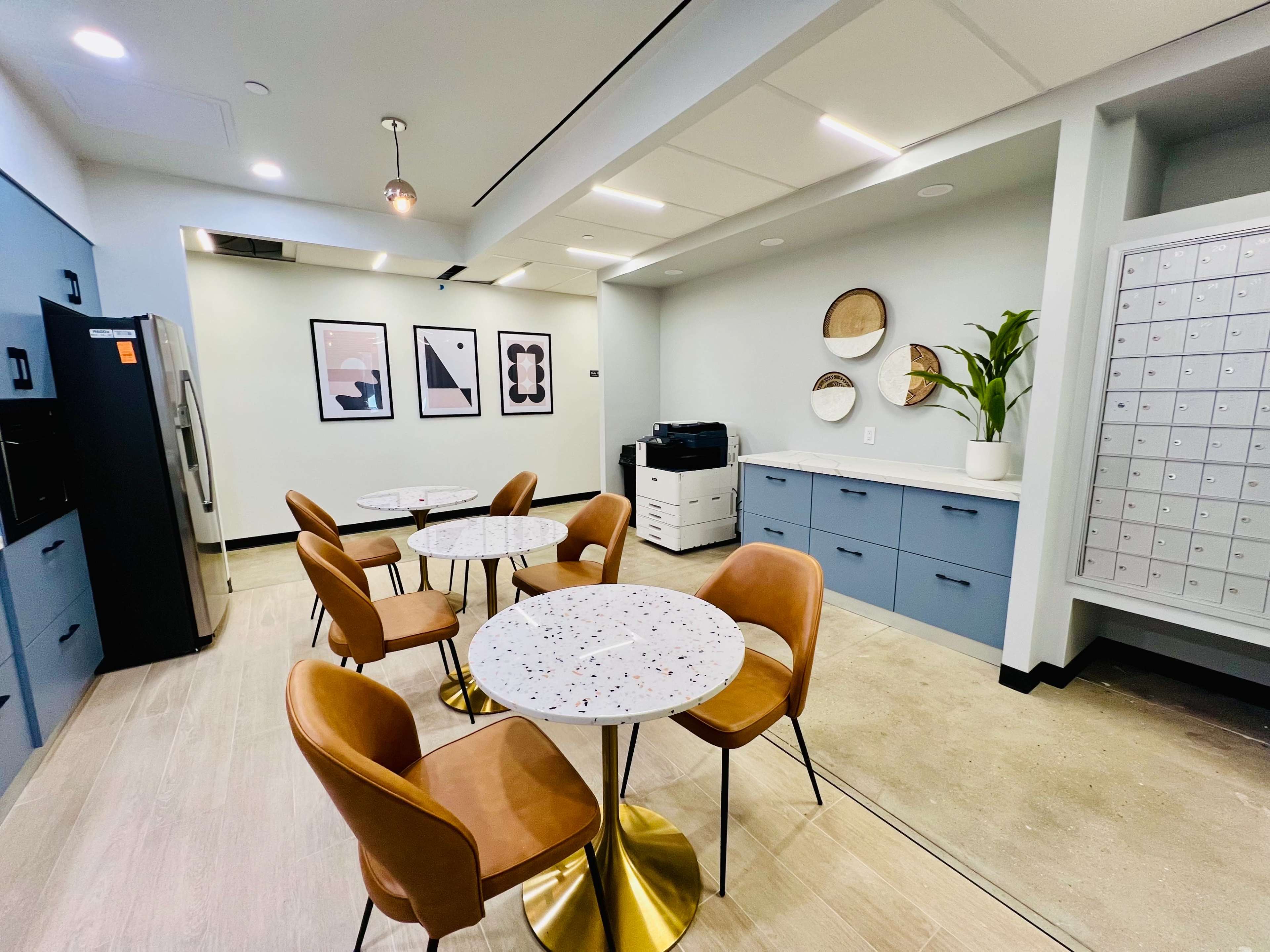 A modern communal space features four circular tables with brown chairs, a kitchenette with a refrigerator, a printer, and a wall of mailbox slots.