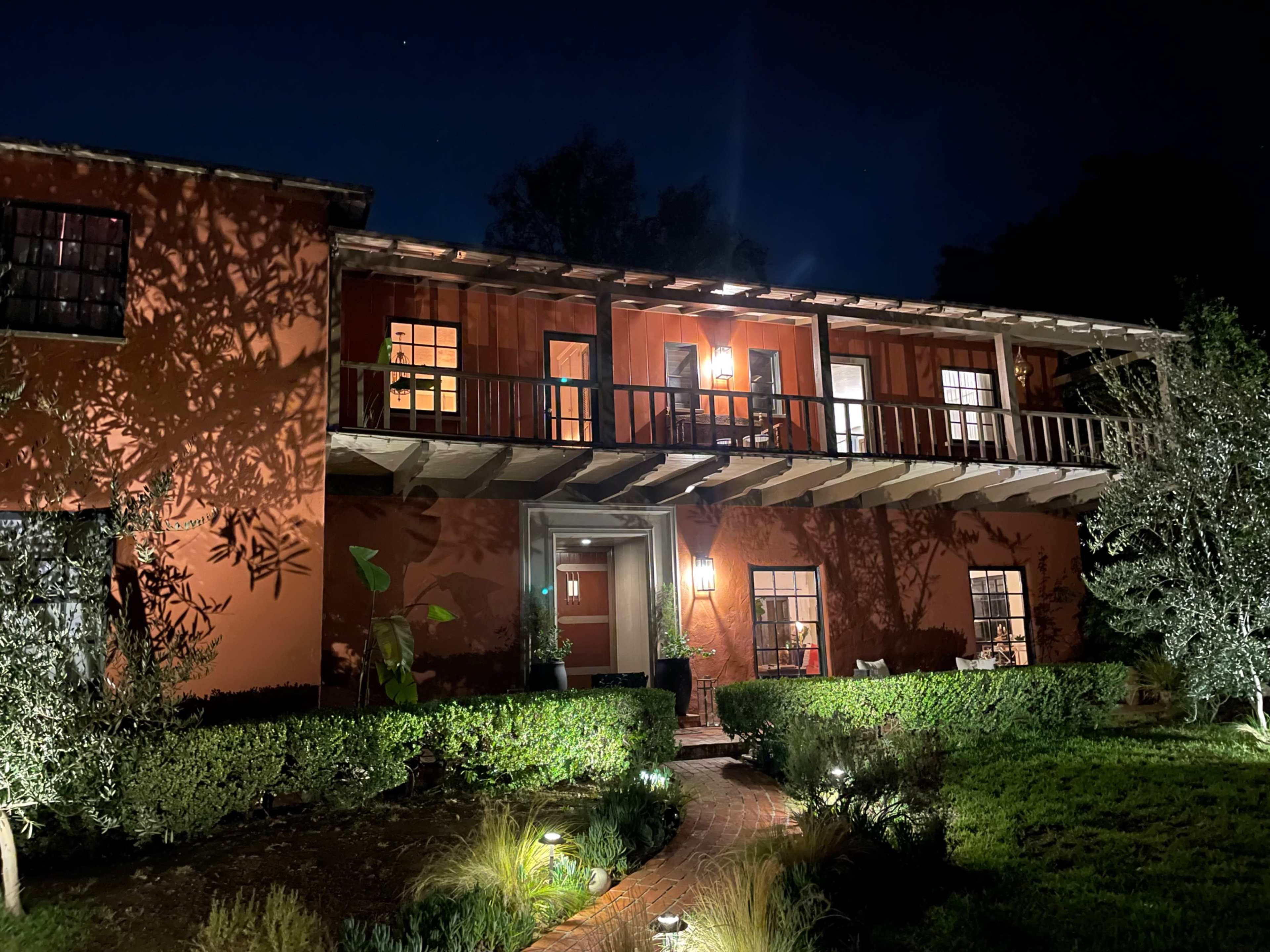 The two-story house features a balcony, illuminated windows, and surrounding greenery, set against a night sky.