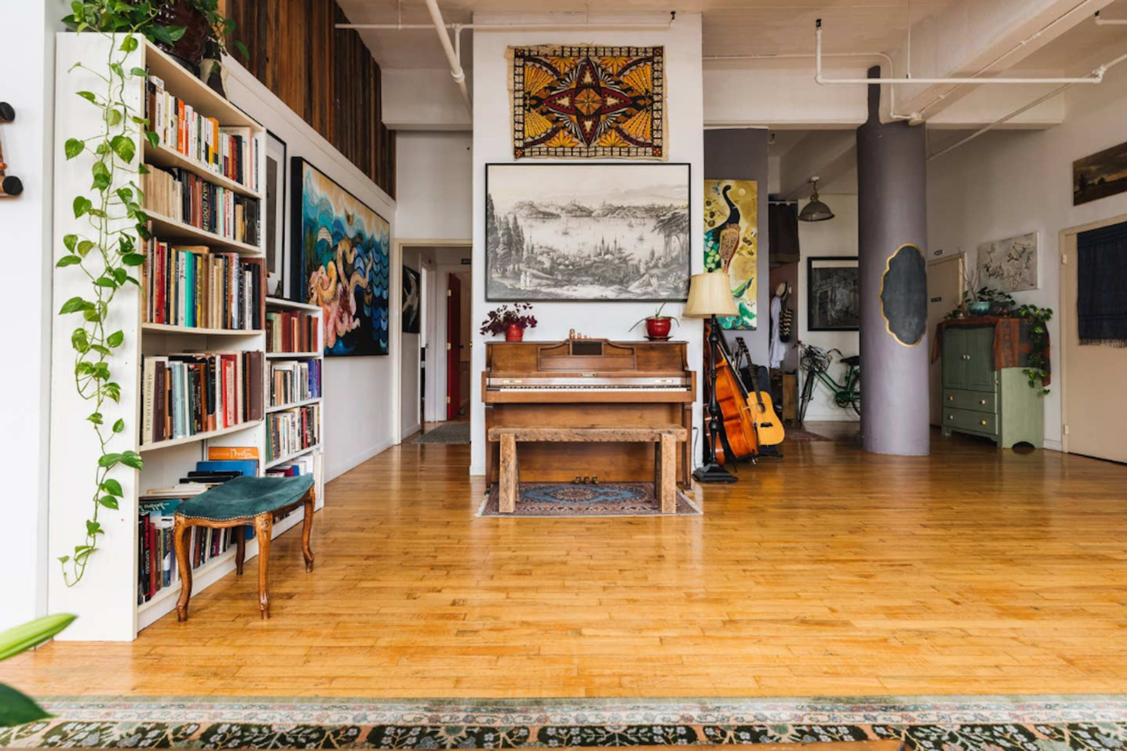 The image shows a spacious room with hardwood floors, featuring a piano, bookshelves filled with books, and various artistic decorations on the walls.