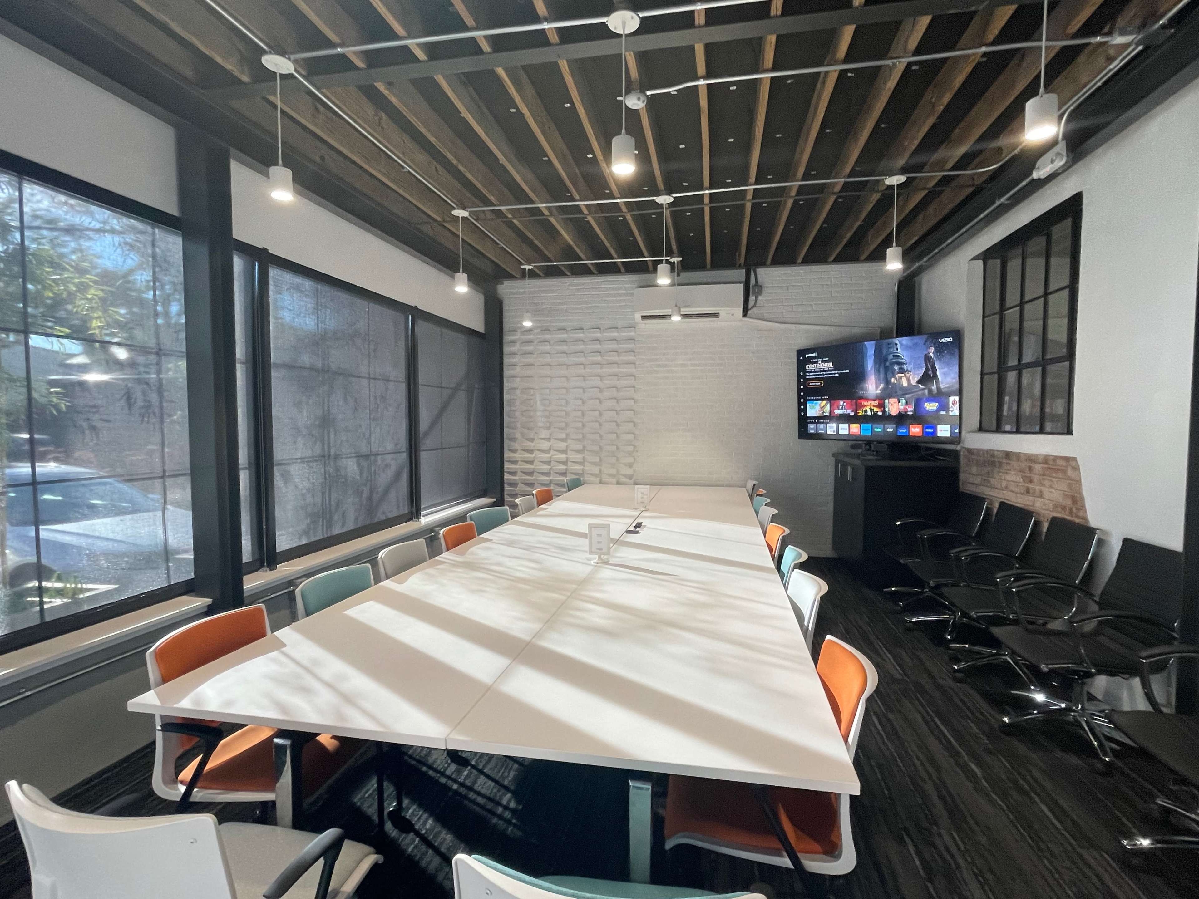 A modern conference room features a long white table surrounded by colorful chairs and a large wall-mounted screen.