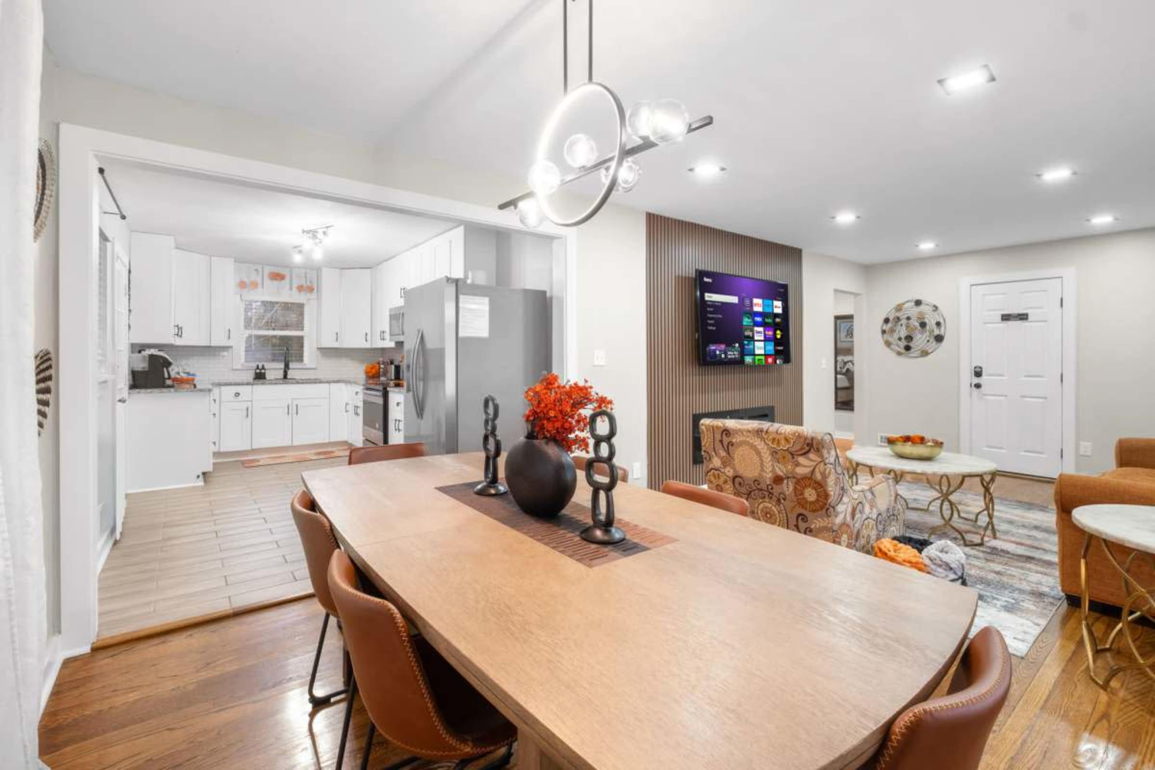 A dining area with a wooden table and modern light fixtures is adjacent to a kitchen with white cabinets and a living room featuring a wall-mounted TV.