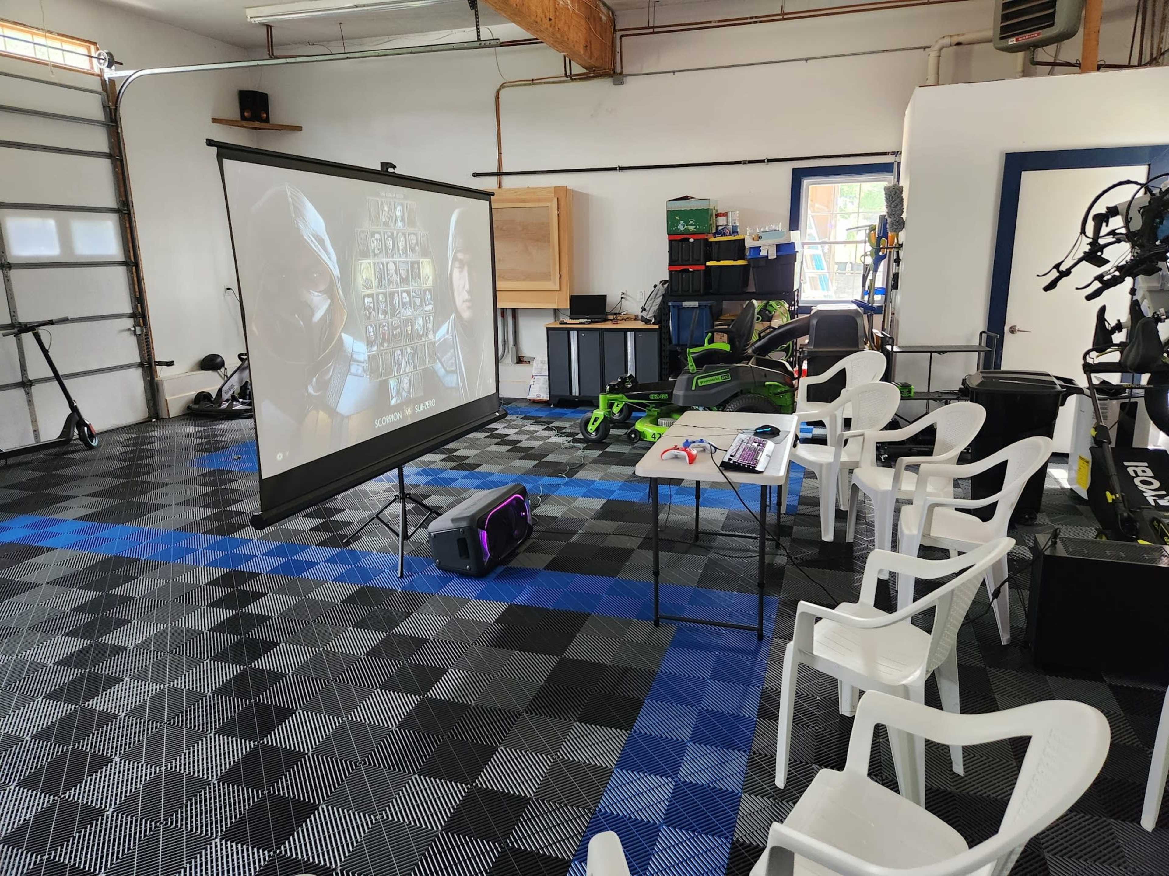 A garage converted into a gaming area, featuring a projector screen displaying characters, a table with gaming equipment, and several white plastic chairs arranged nearby on a checkered floor.