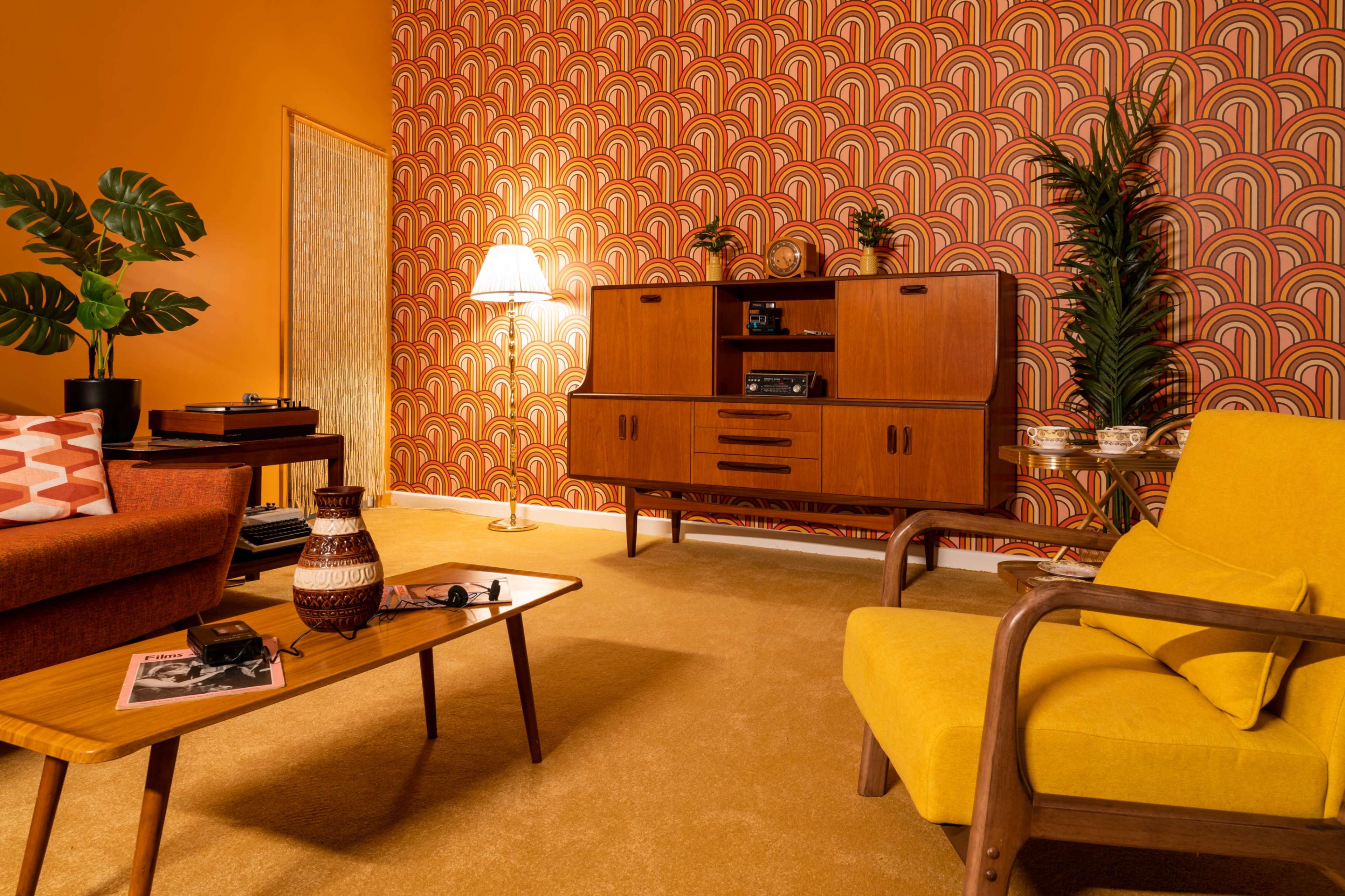 The image shows a retro-style living room with orange walls patterned in arcs, featuring wooden furniture, a yellow armchair, and a coffee table.