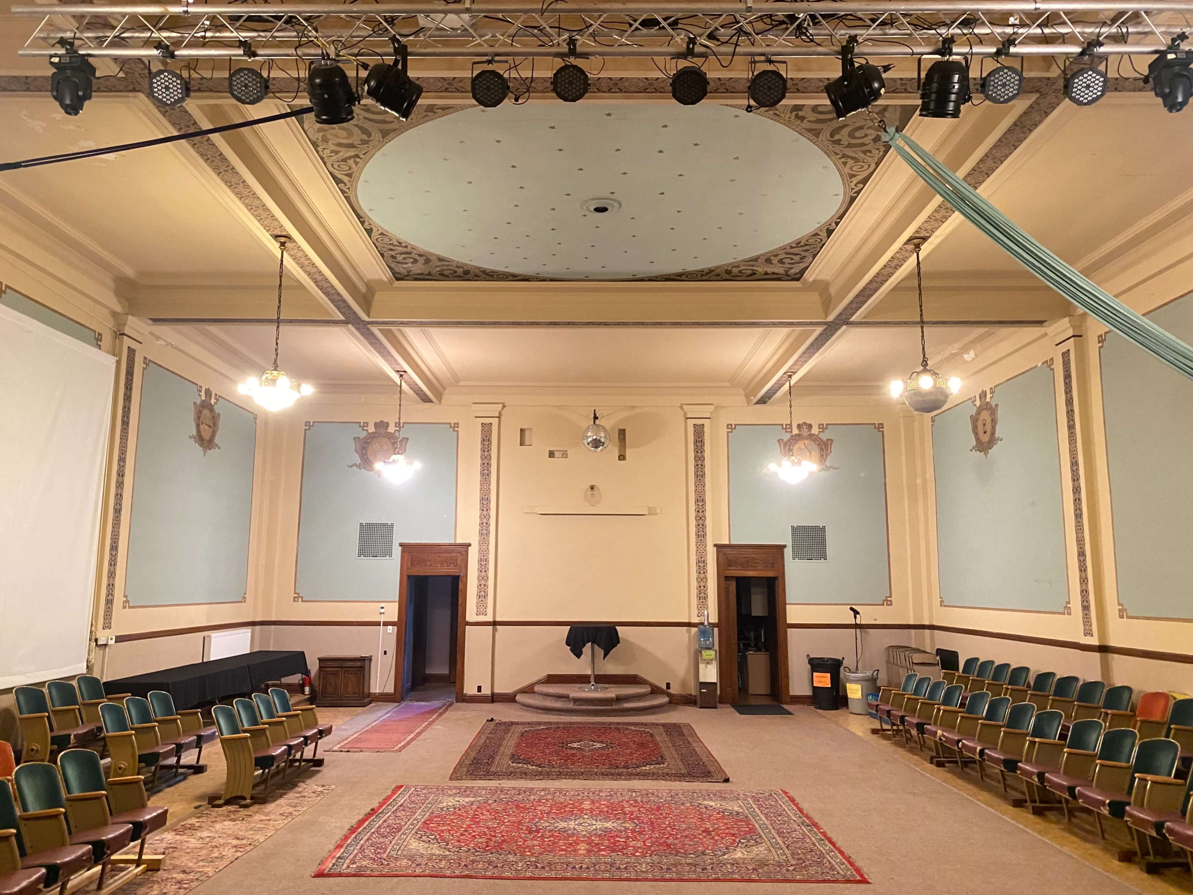A spacious hall with rows of green chairs, a large decorative ceiling, and a stage at the front, flanked by two open doors.