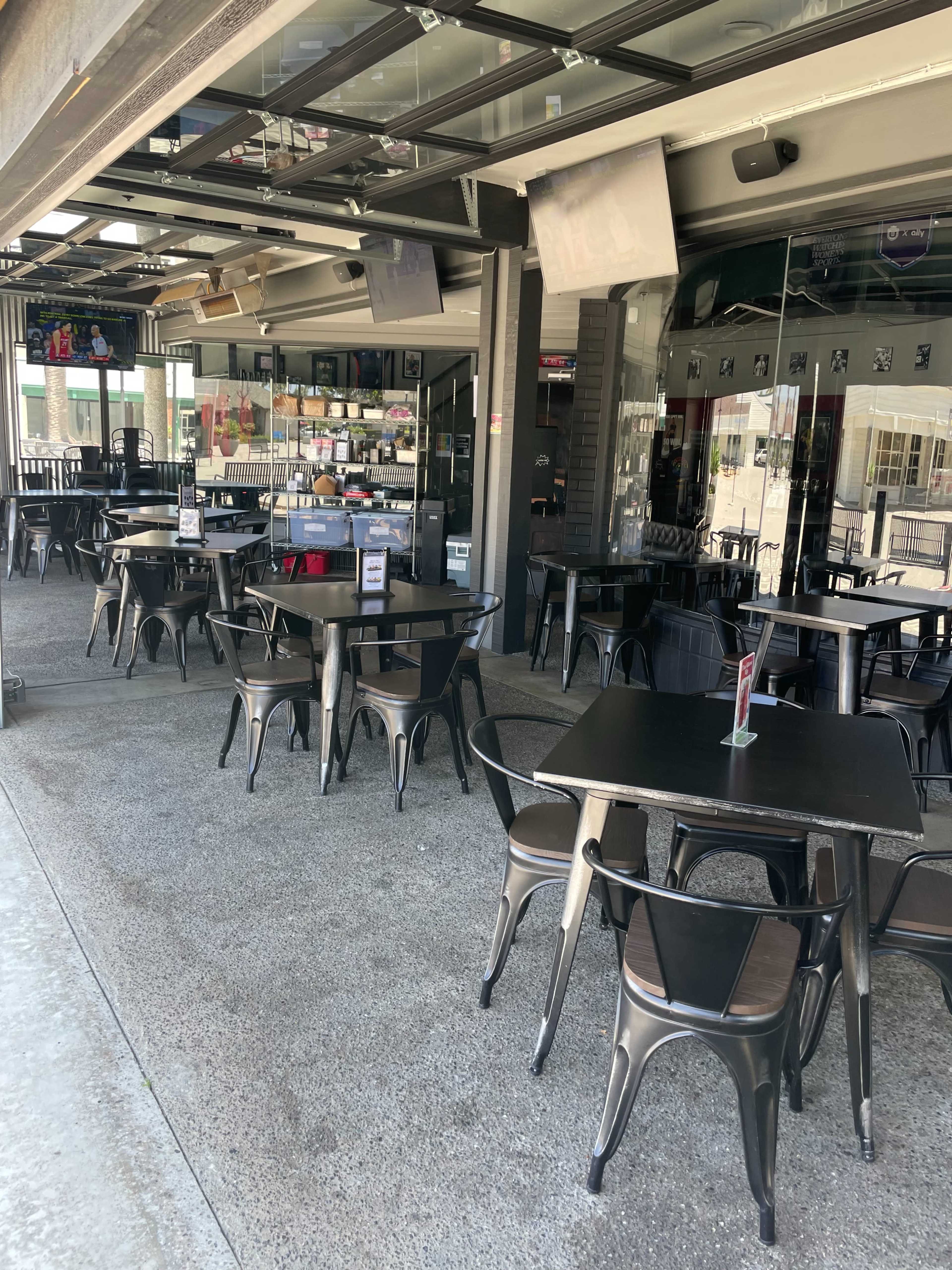 The image shows a modern restaurant with multiple black tables and chairs arranged on a concrete floor, featuring large glass windows and a partially open entrance.