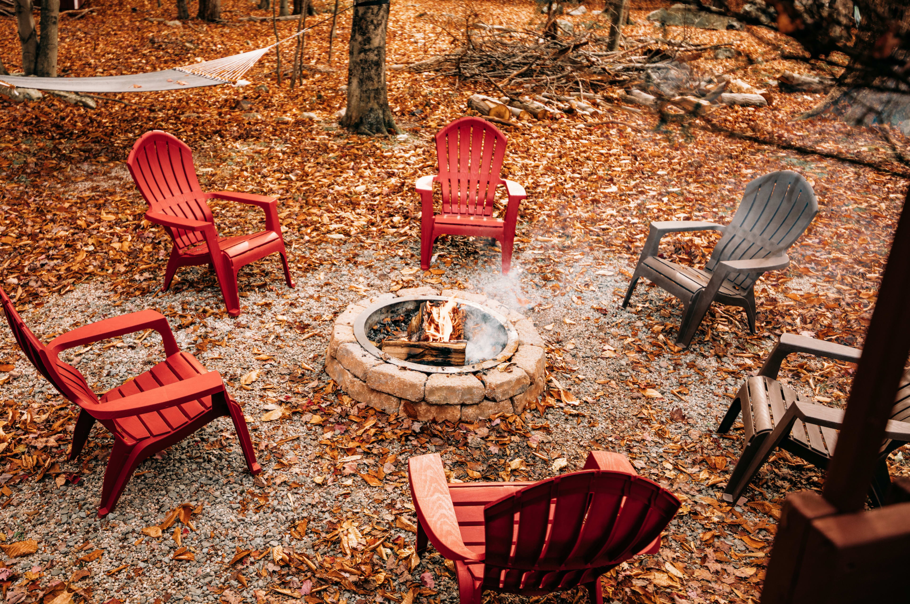 A stone fire pit surrounded by six adirondack chairs on a bed of fallen leaves in a wooded area.