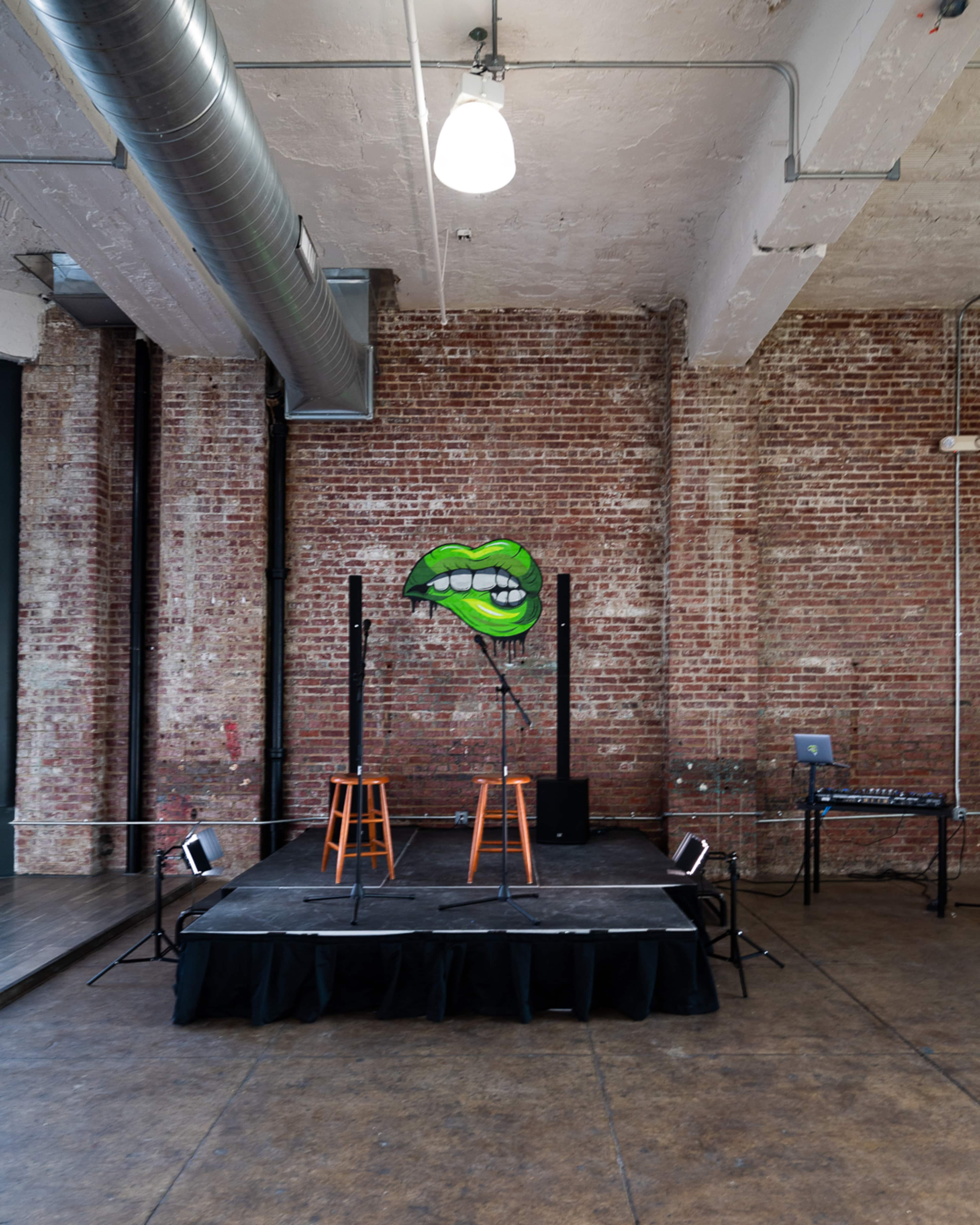 A stage with two wooden stools, a microphone stand adorned with a large green lips design, and a music setup in the background within a brick-walled room.
