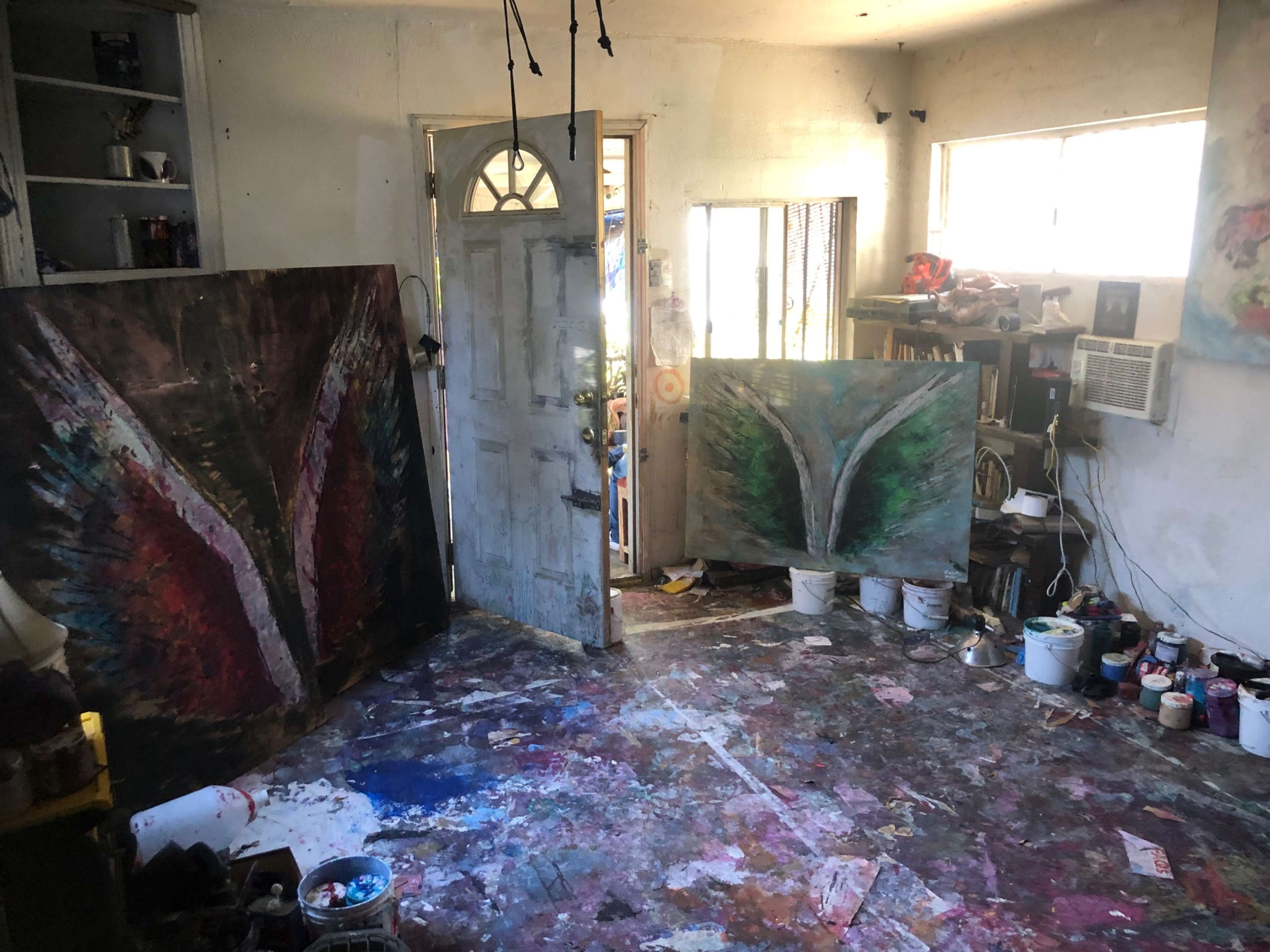 The image shows an artist's studio with two large paintings leaning against the walls, colorful paint splatters on the floor, and an open door leading outside.
