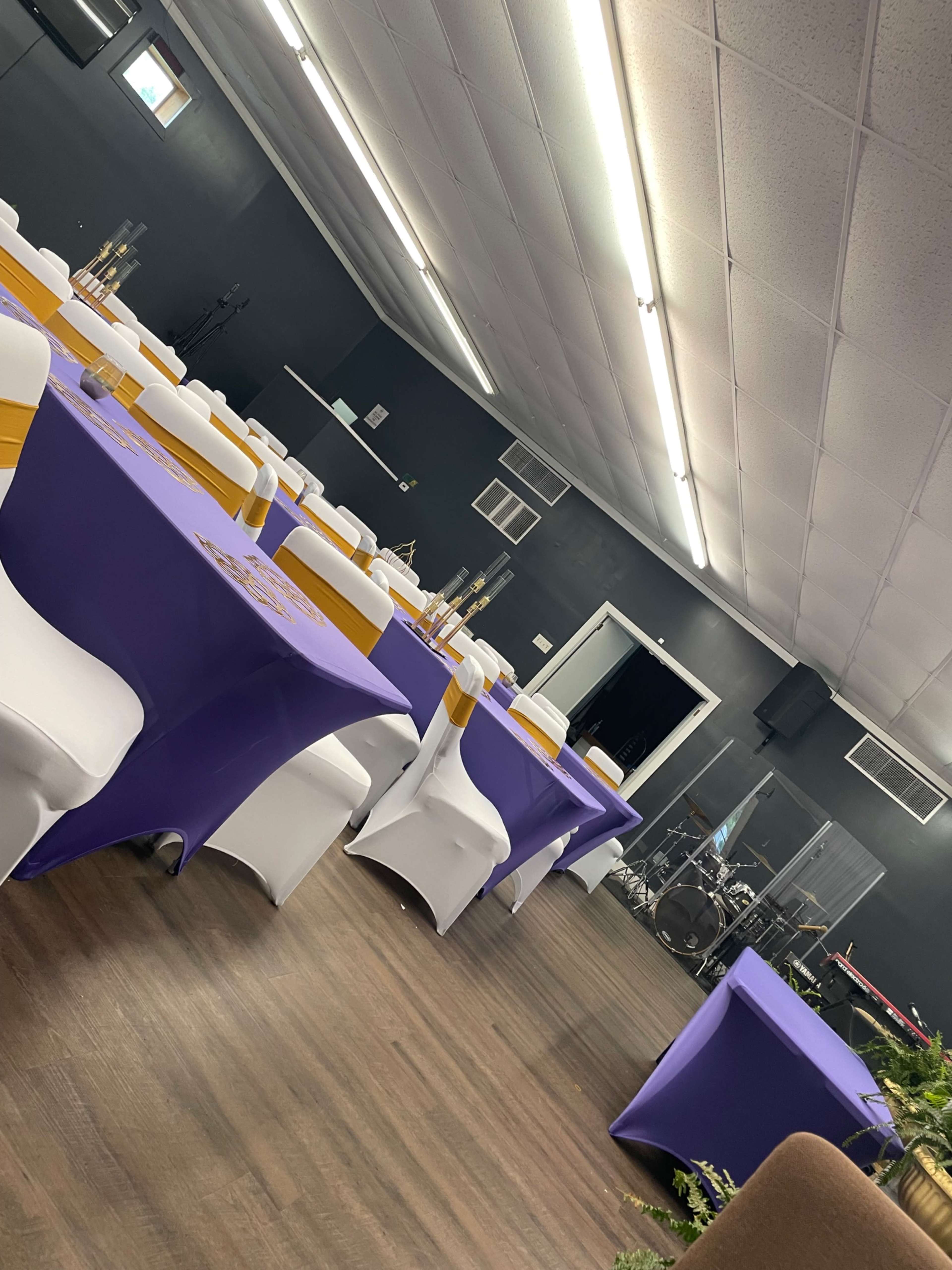 A decorated event space with purple tablecloths and white chairs arranged in rows, featuring a stage area in the background.