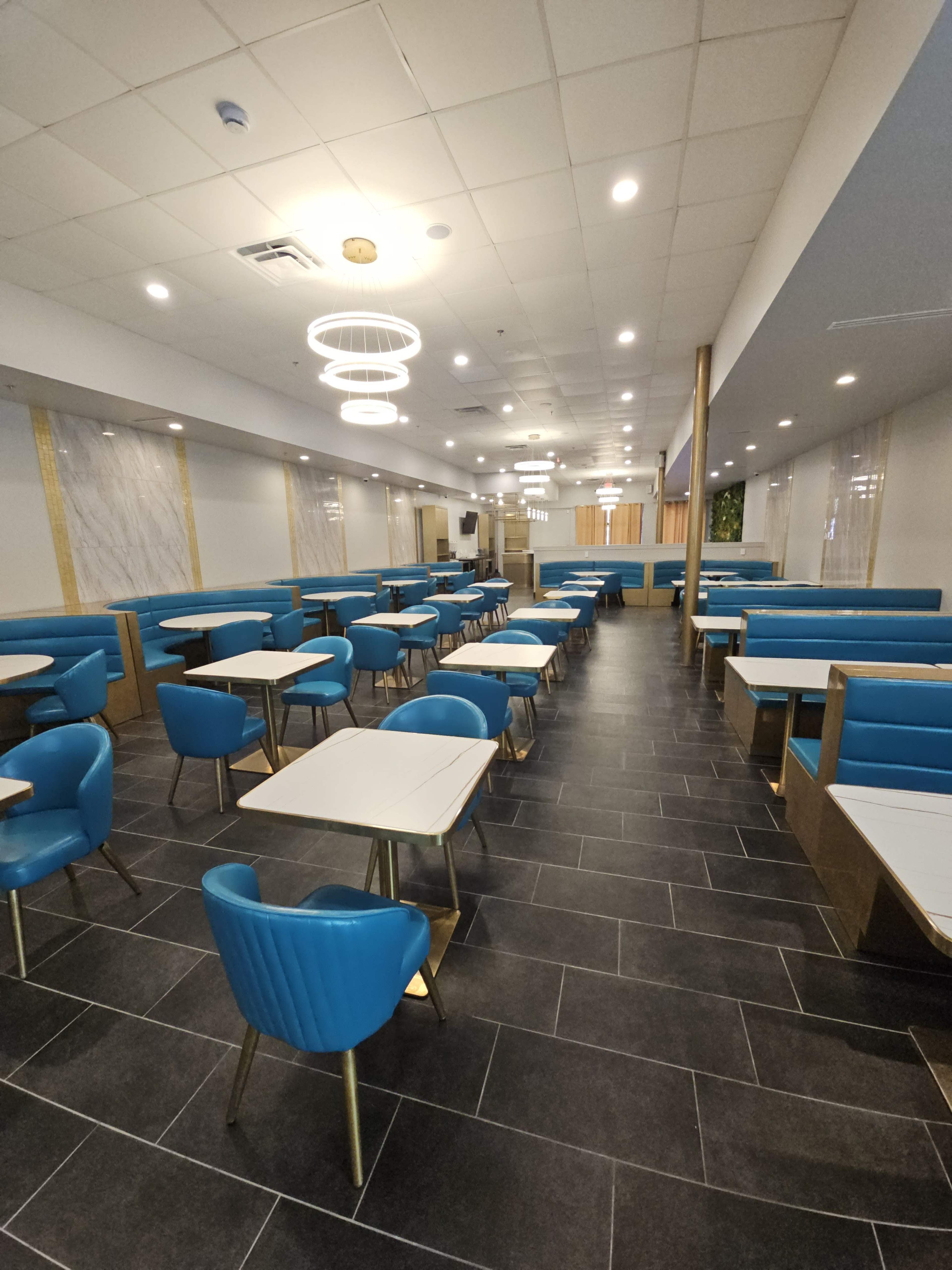 The image shows an interior of a restaurant with rows of blue chairs and tables arranged neatly across a dark tiled floor.