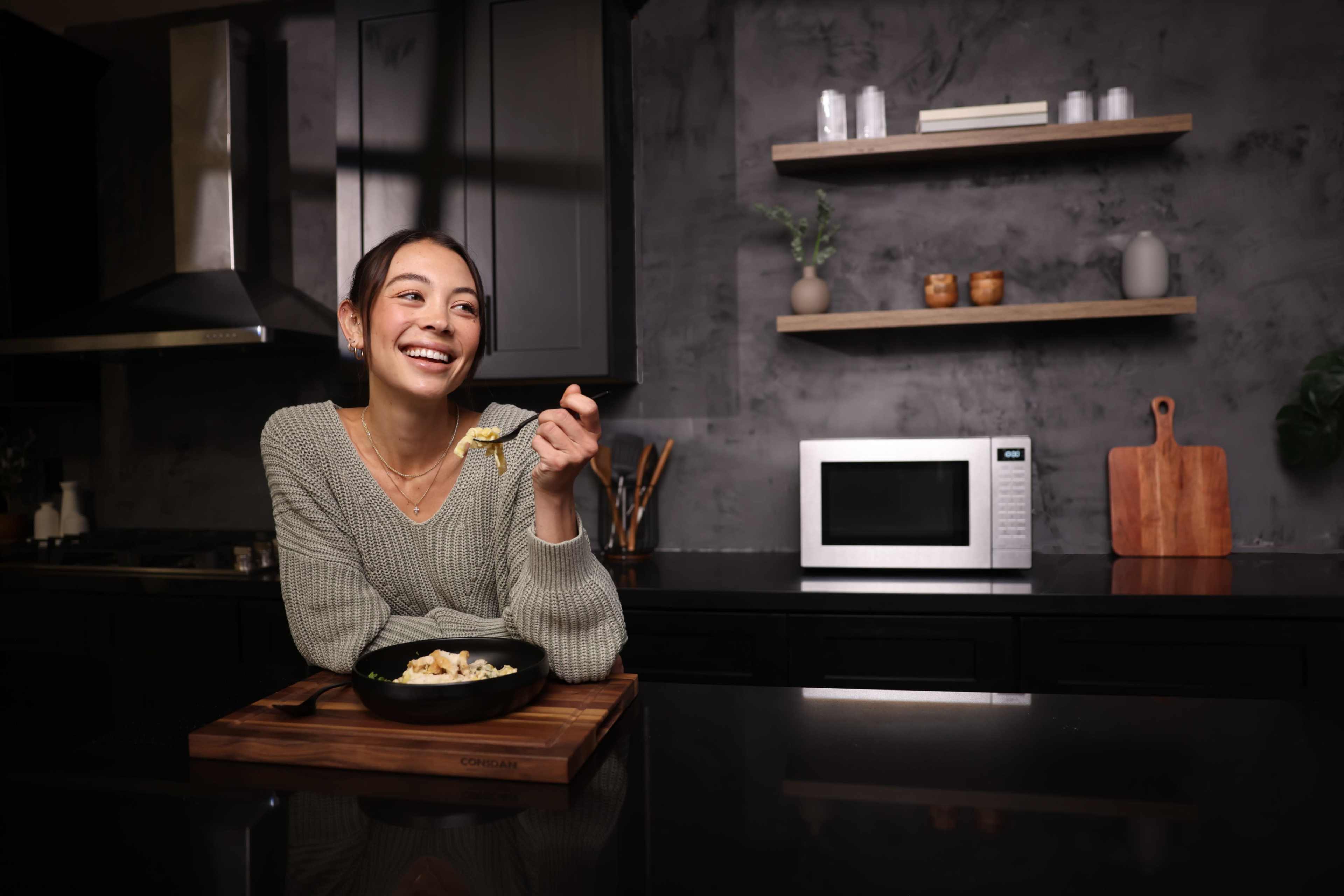 A woman sits at a kitchen island, smiling while holding a forkful of pasta, with a microwave and shelf of kitchen items in the background.