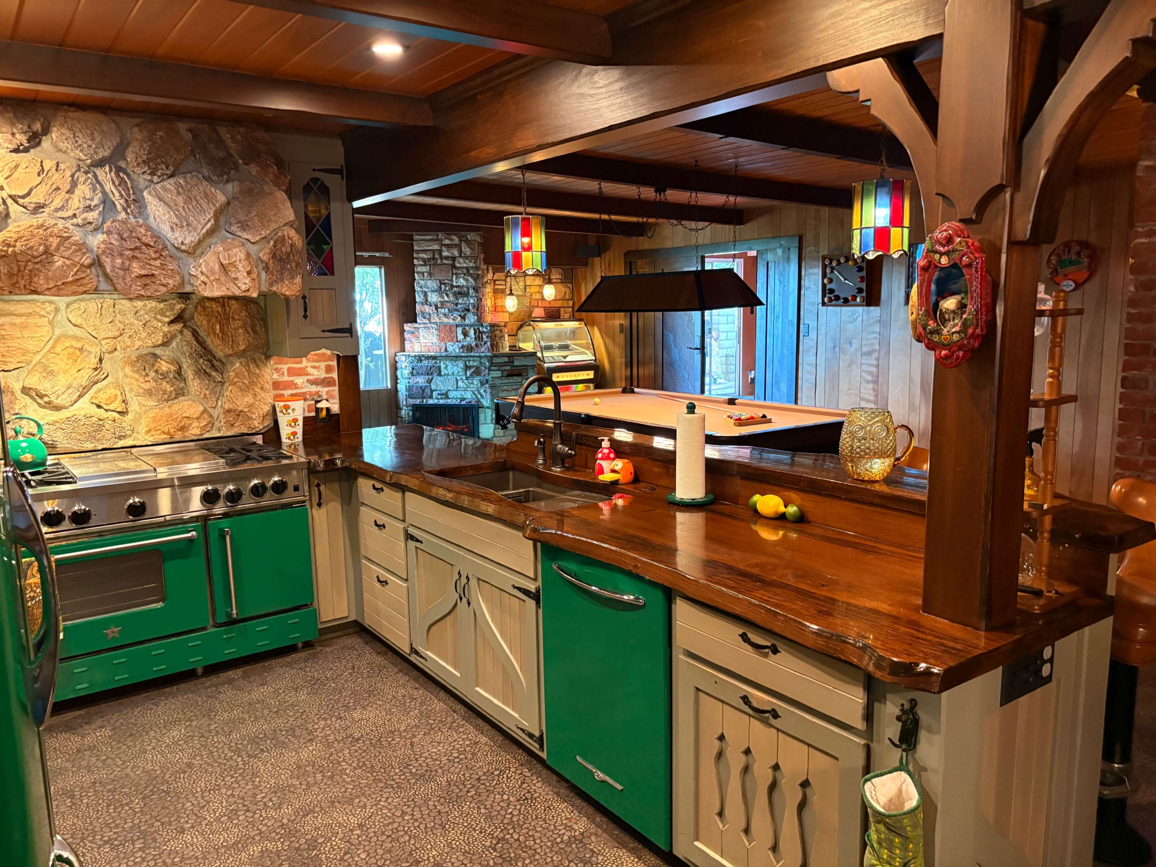 The image shows a kitchen featuring green cabinets, a stone wall, wooden beams, and a large island with a countertop and various kitchen appliances.