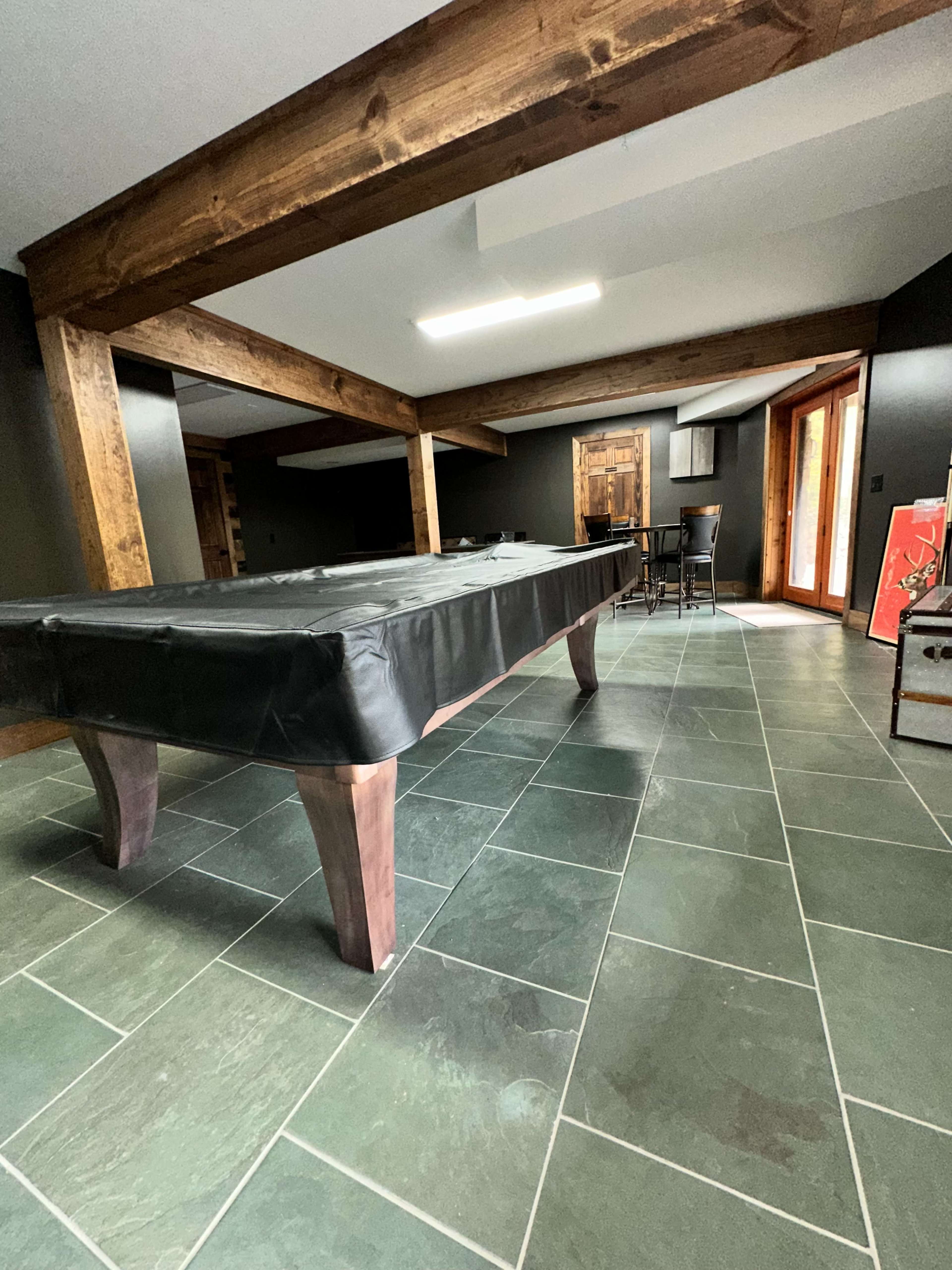 The image shows a game room featuring a black-covered pool table surrounded by slate flooring and wooden beams.