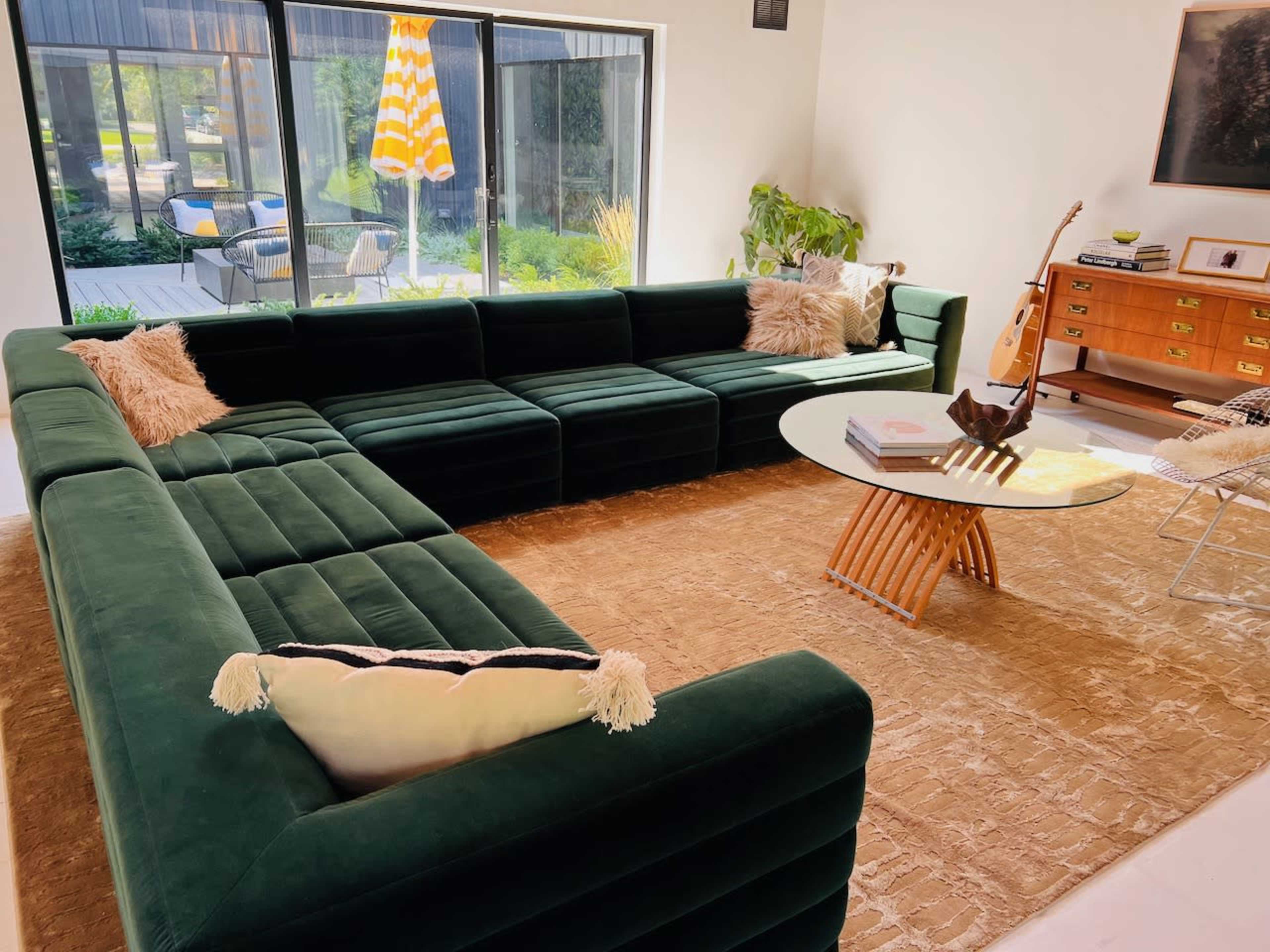 A spacious living room features a large green sectional sofa, a round glass table, and a wooden cabinet, with large windows overlooking an outdoor patio.