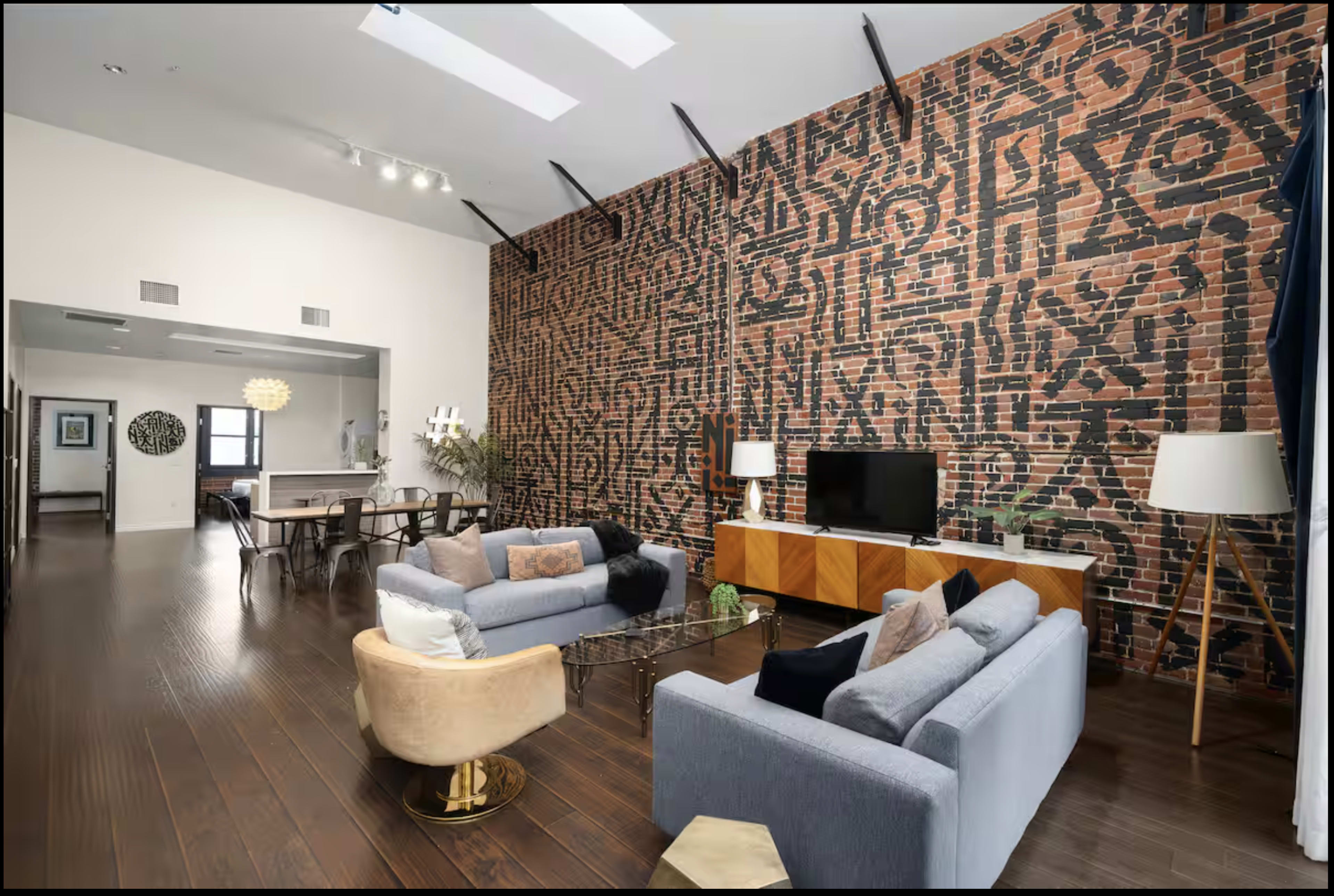 The image shows a modern living room with a brick wall featuring black abstract patterns, two sofas, a coffee table, and a television on a wooden media console.