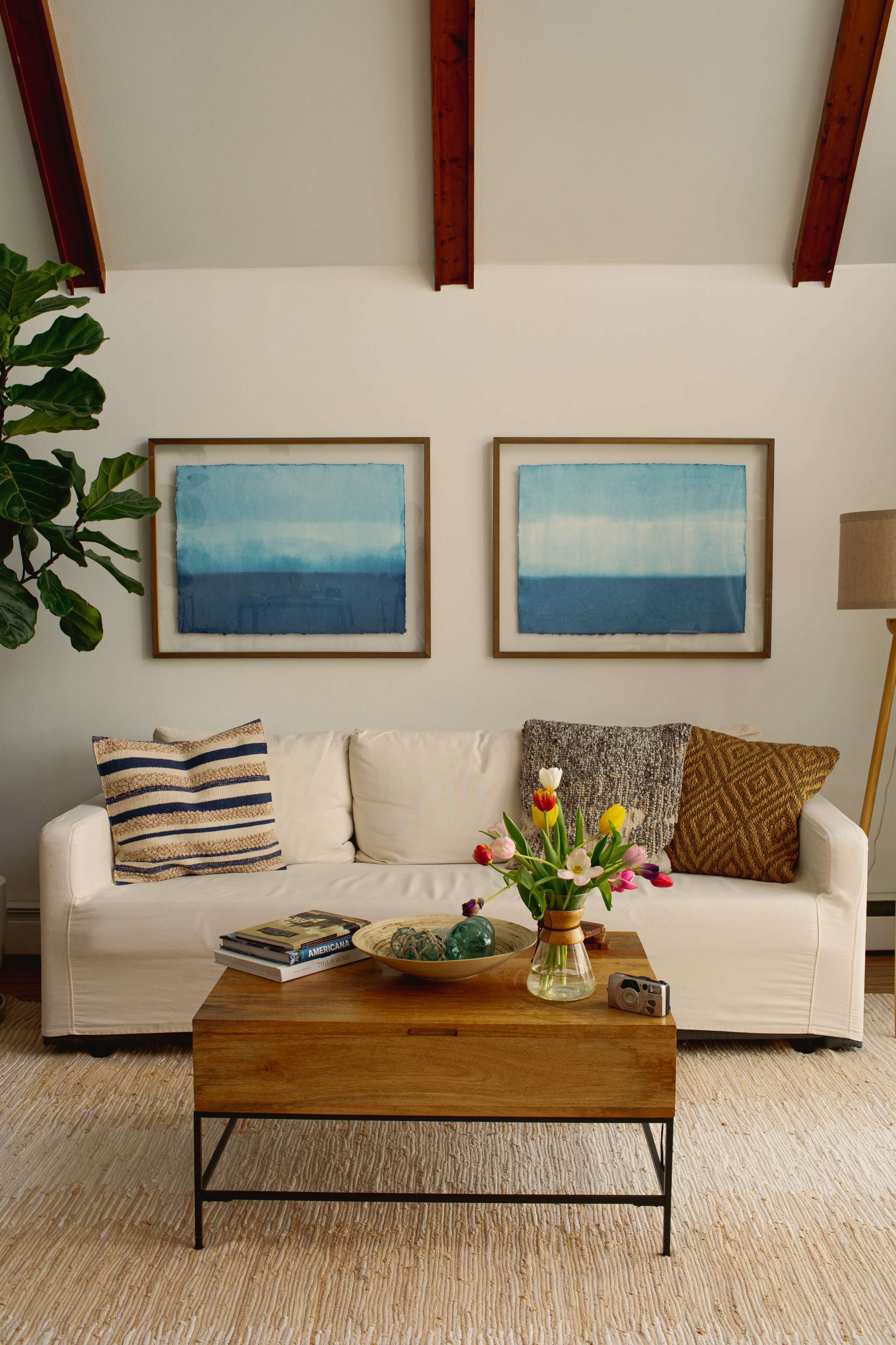 A cozy living room features a white sofa with decorative pillows, a wooden coffee table with books and decor, and two framed blue gradient artworks on the walls.