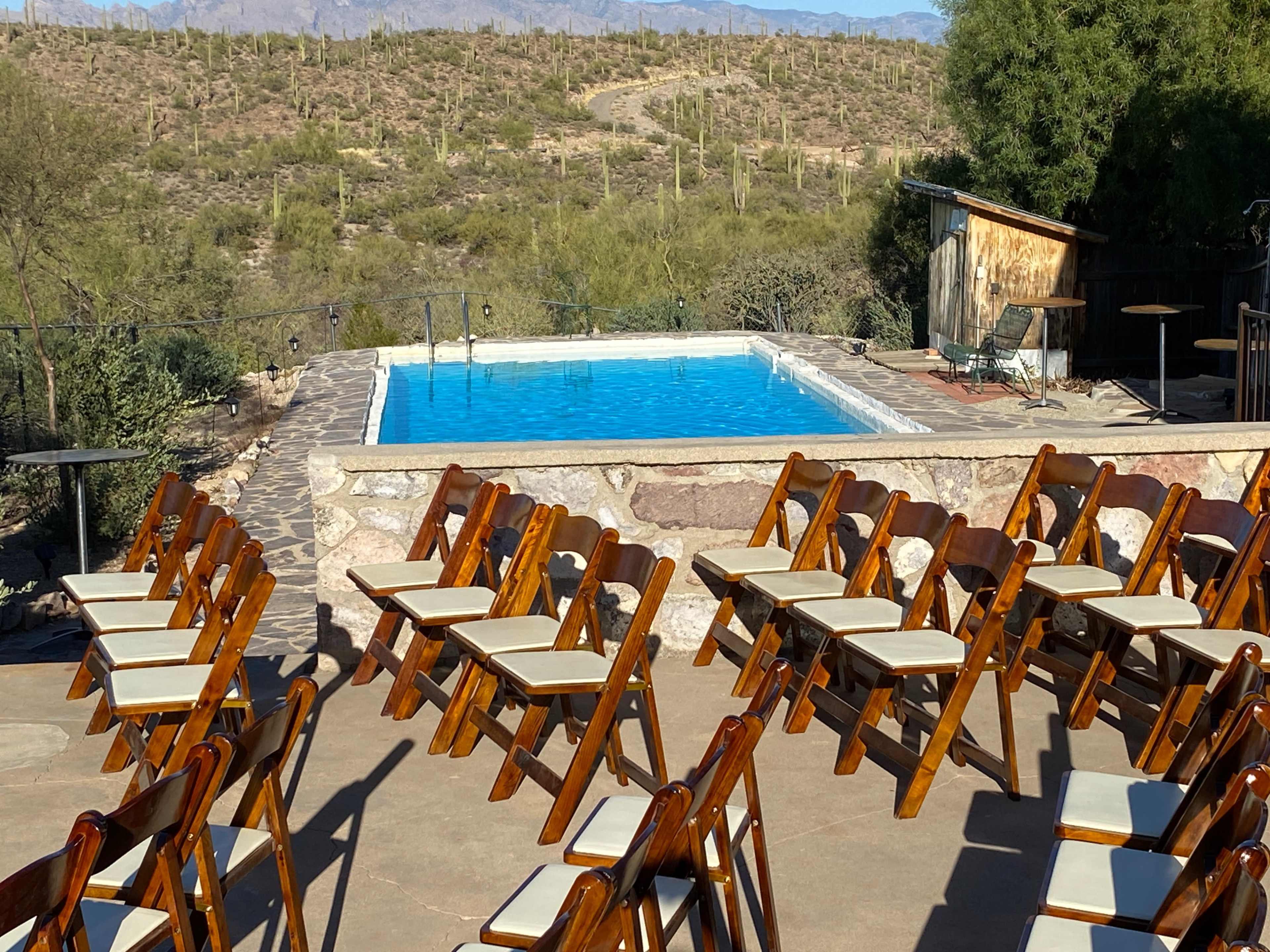 The image shows a set of wooden chairs arranged around a swimming pool, with a desert landscape and mountains in the background.