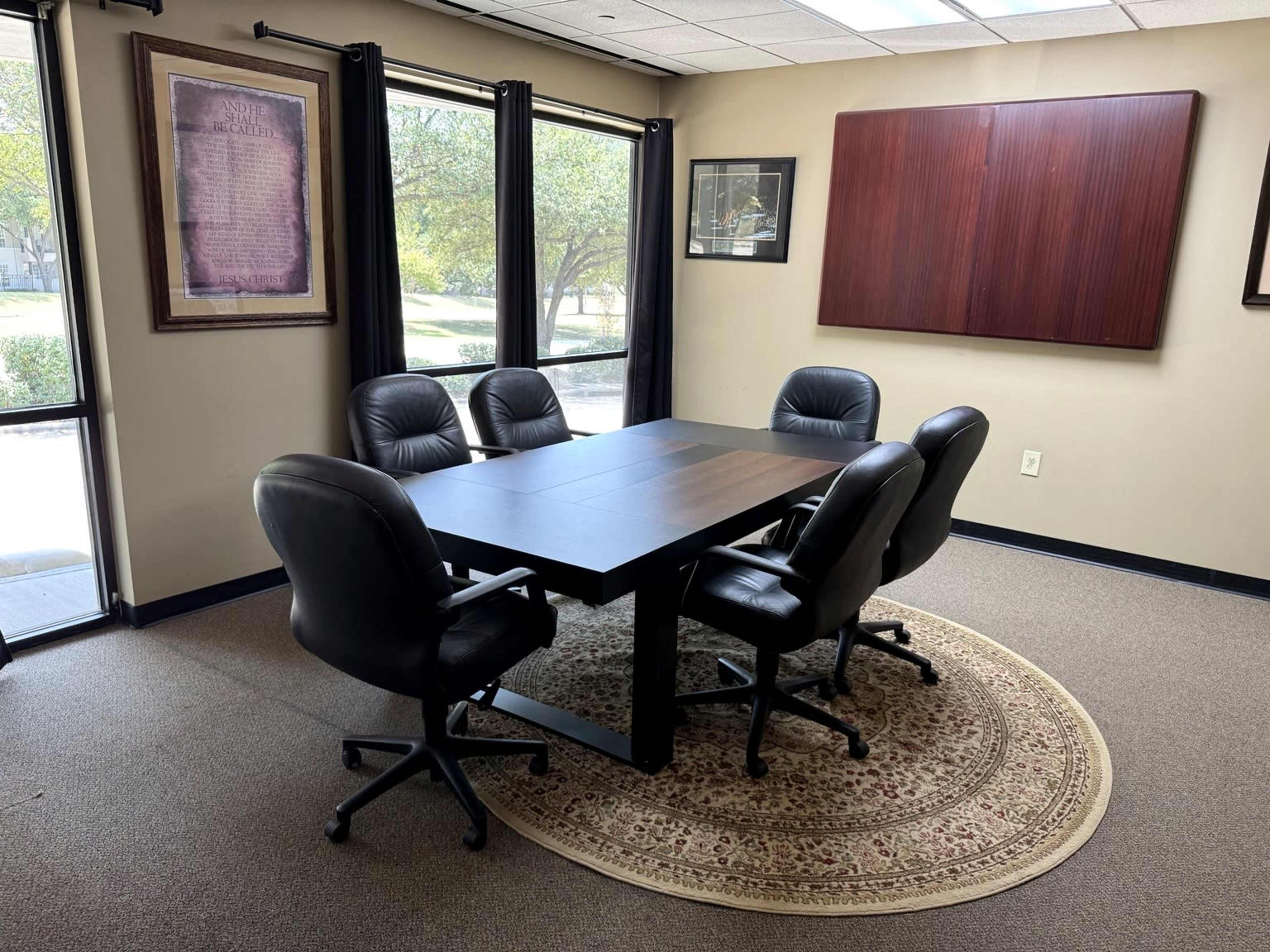 The image shows a conference room with a rectangular table surrounded by six black leather chairs on a patterned area rug, with large windows letting in natural light.