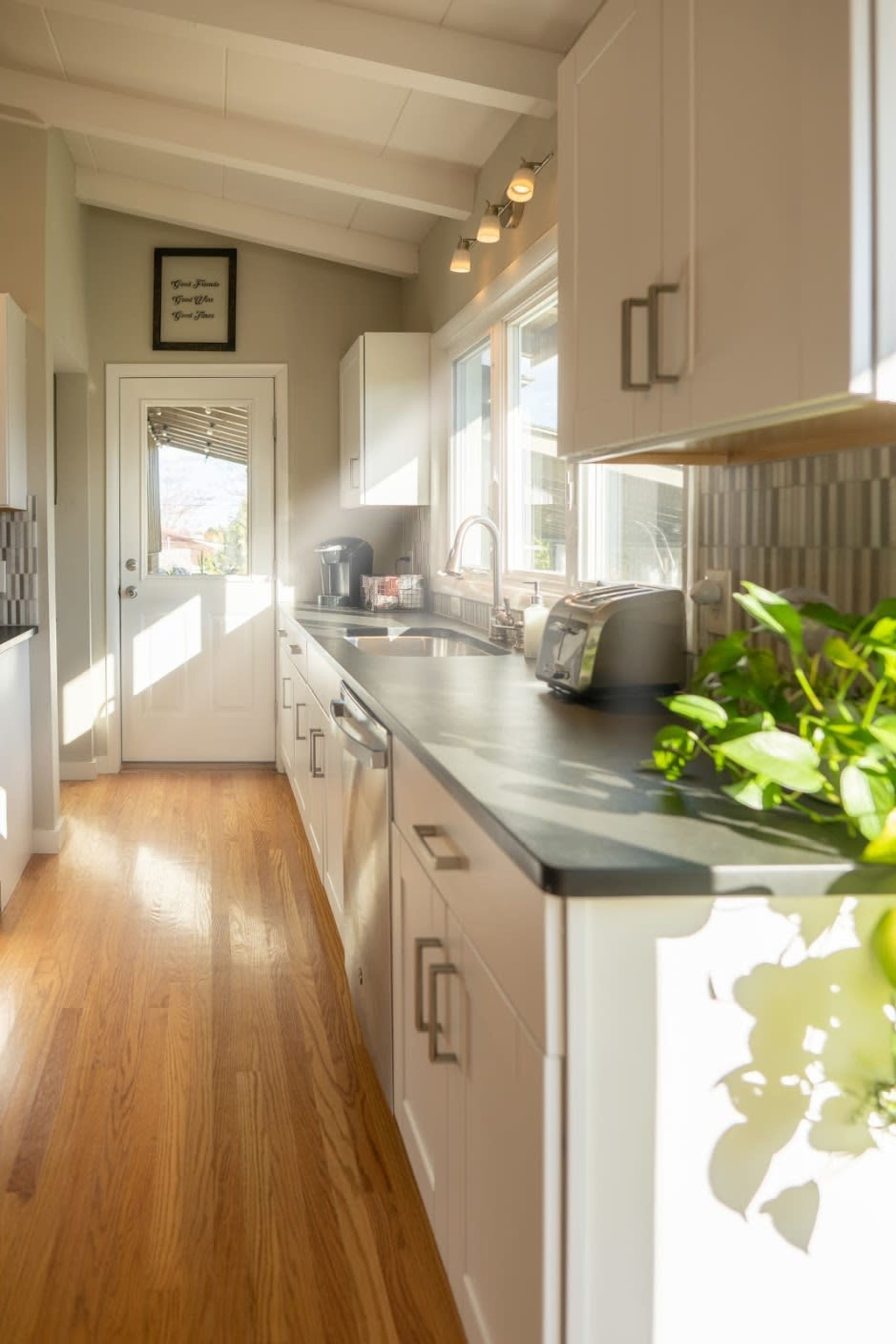 The kitchen features white cabinetry, a countertop, and a door leading outside, illuminated by natural light.