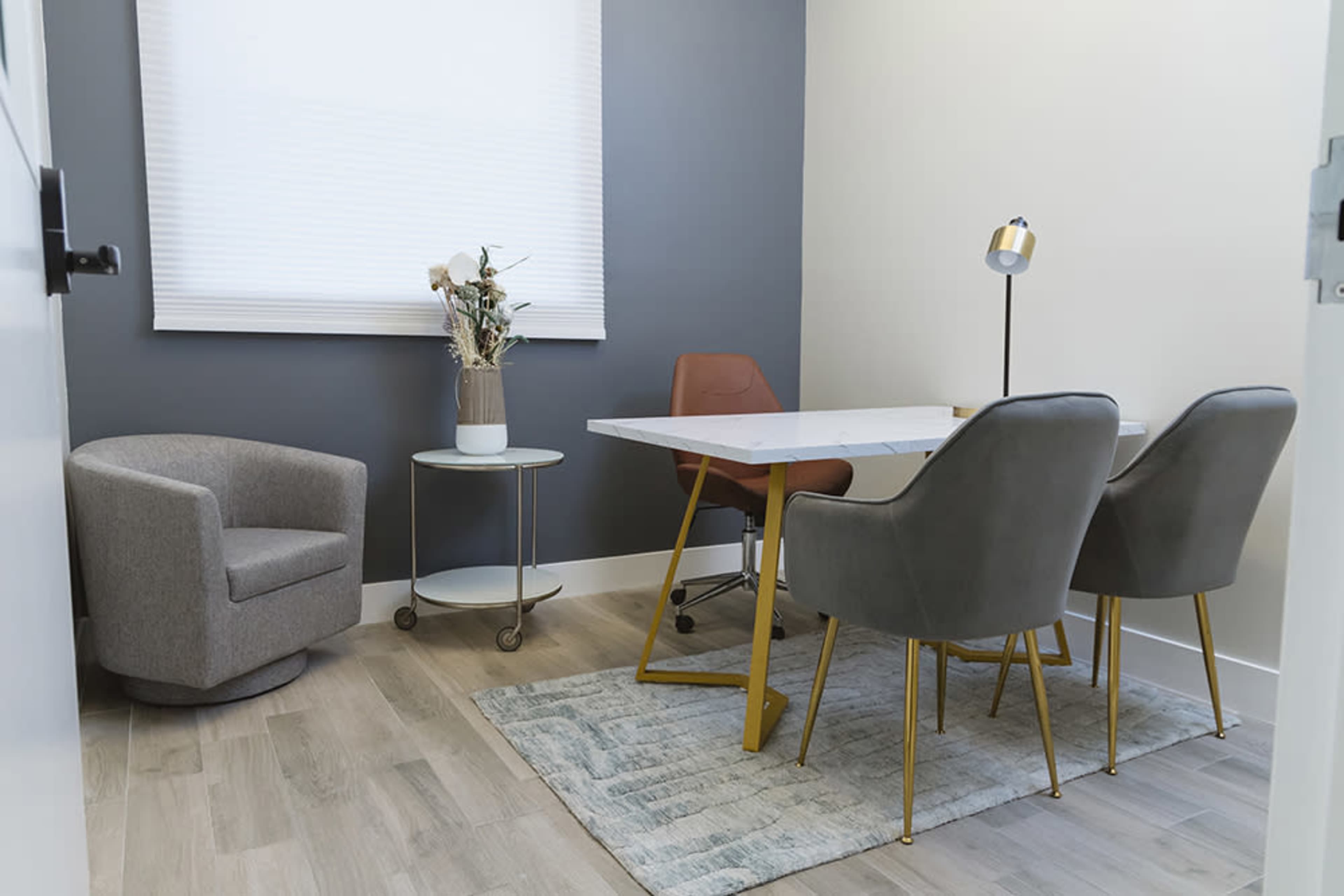 A small office with a round gray chair, a white table with gold legs, two gray chairs, a lamp, and a small side table featuring a vase with dried flowers.