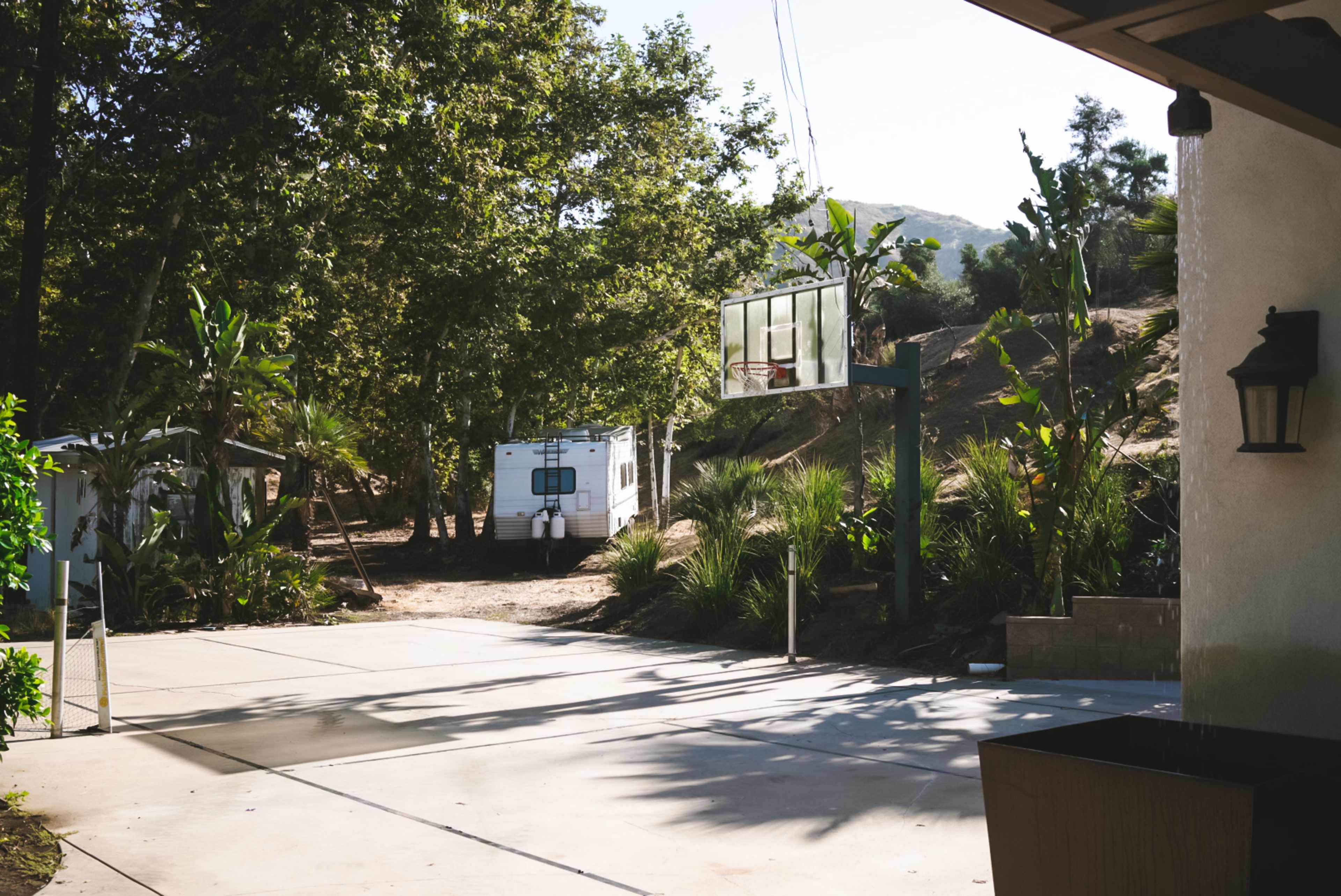 A driveway with a basketball hoop on one side and a white recreational vehicle parked in the background, surrounded by greenery and hills.
