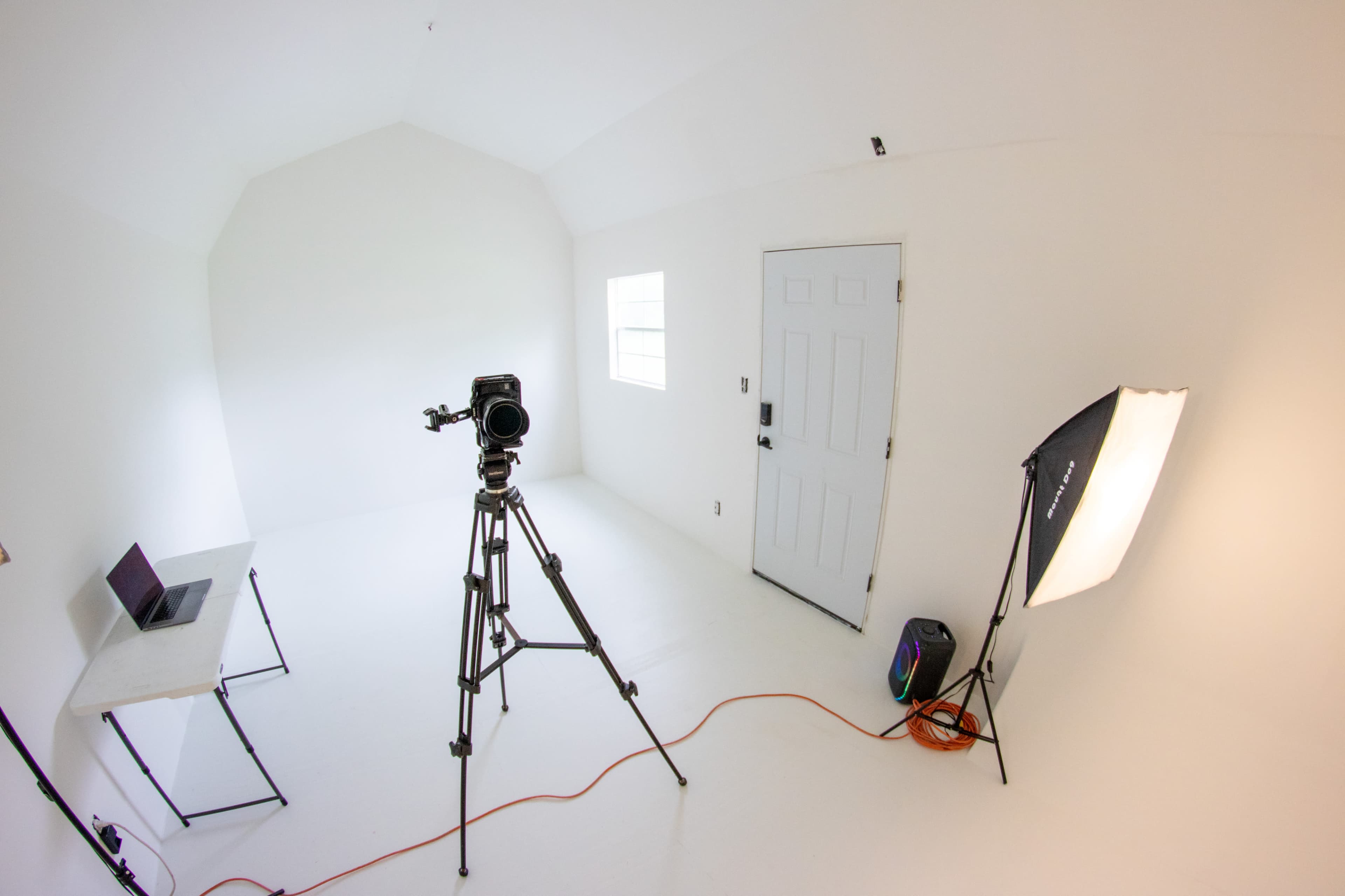 The image shows a small, empty room with white walls, a laptop on a table, a camera on a tripod, and a softbox light near the door.