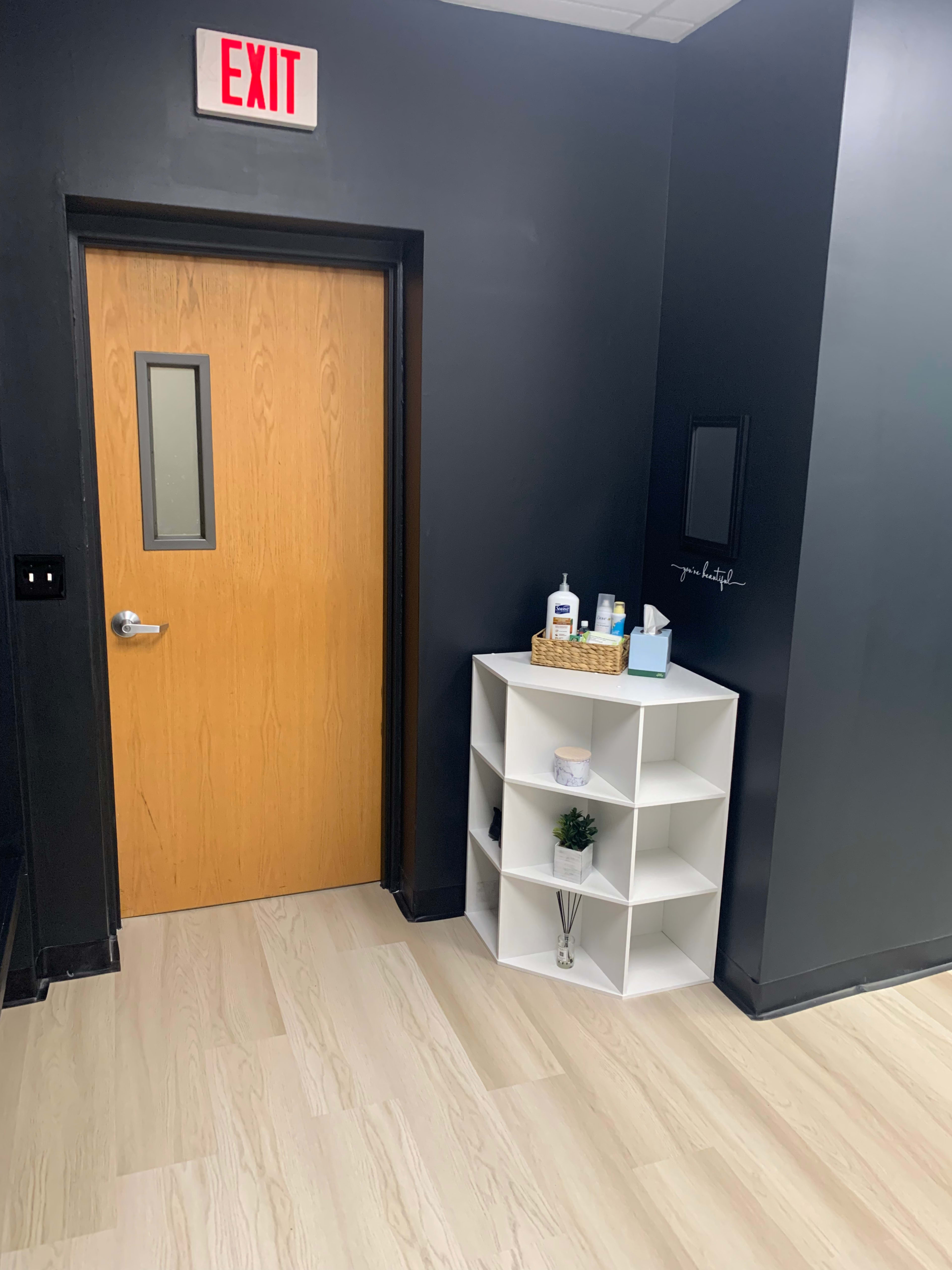 A minimalist interior featuring a wooden exit door, a small white shelving unit with various items, and a dark wall.