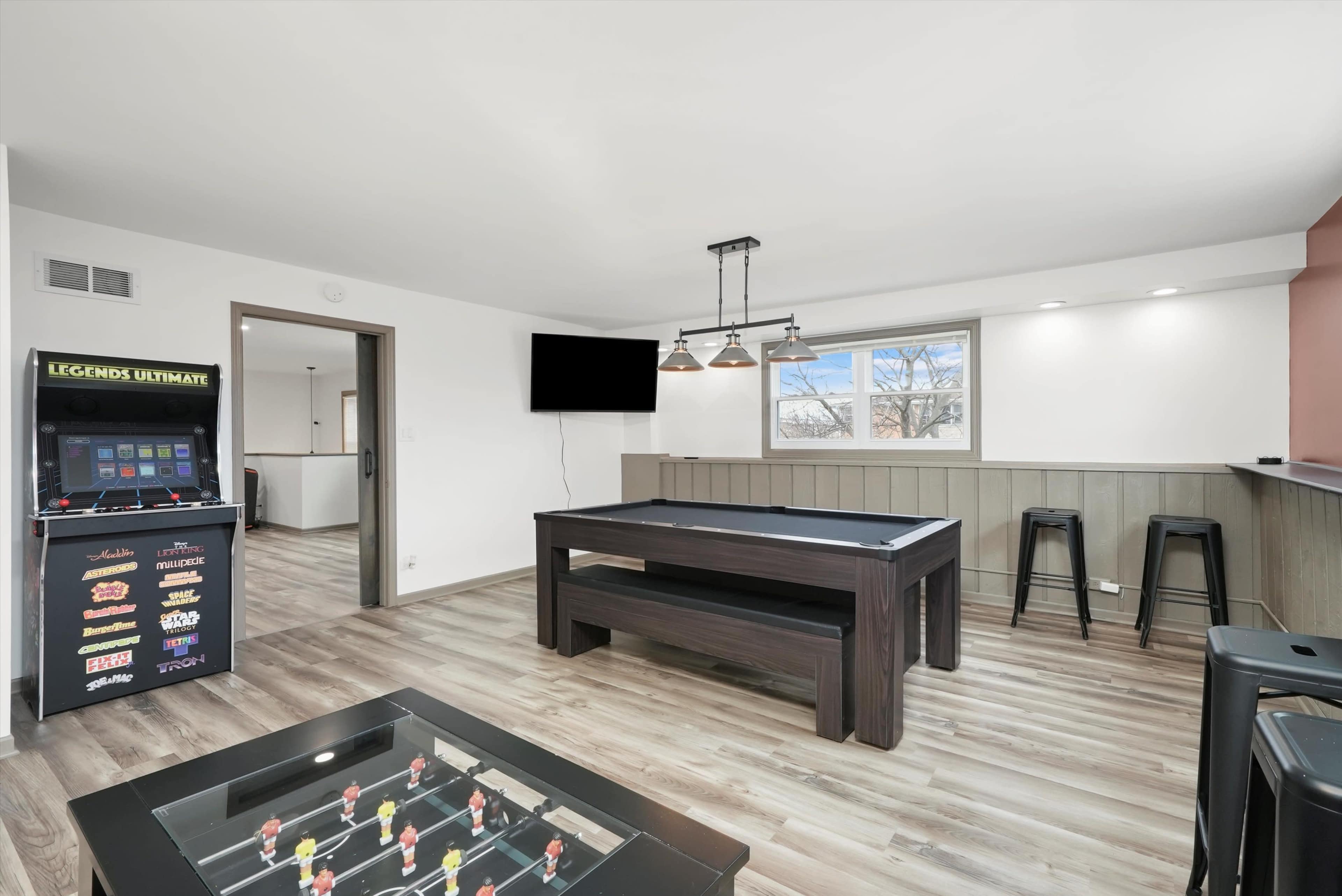 A modern game room featuring a pool table, arcade machine, and bar-height seating near a large window.
