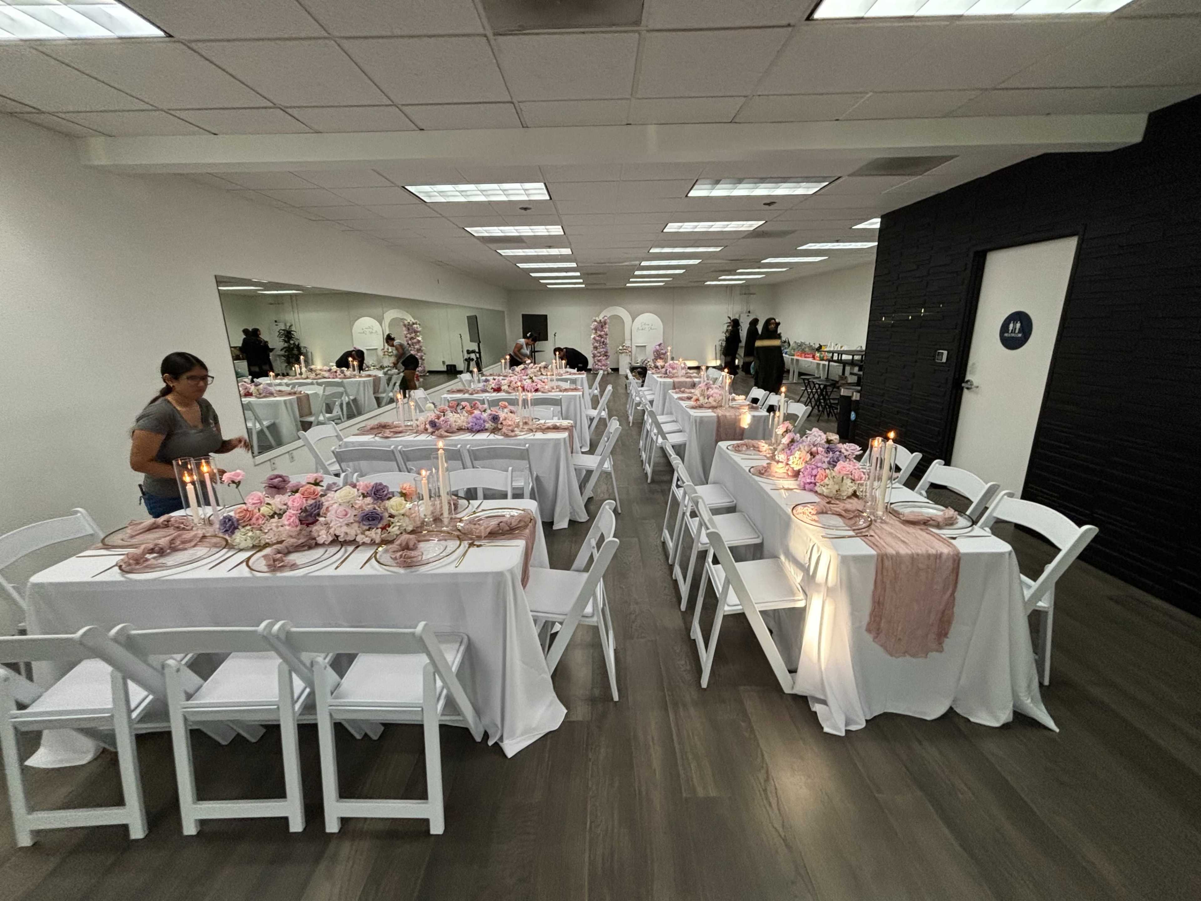 A spacious event room is set with multiple white tables adorned with floral centerpieces and candles, while a person prepares for the gathering.
