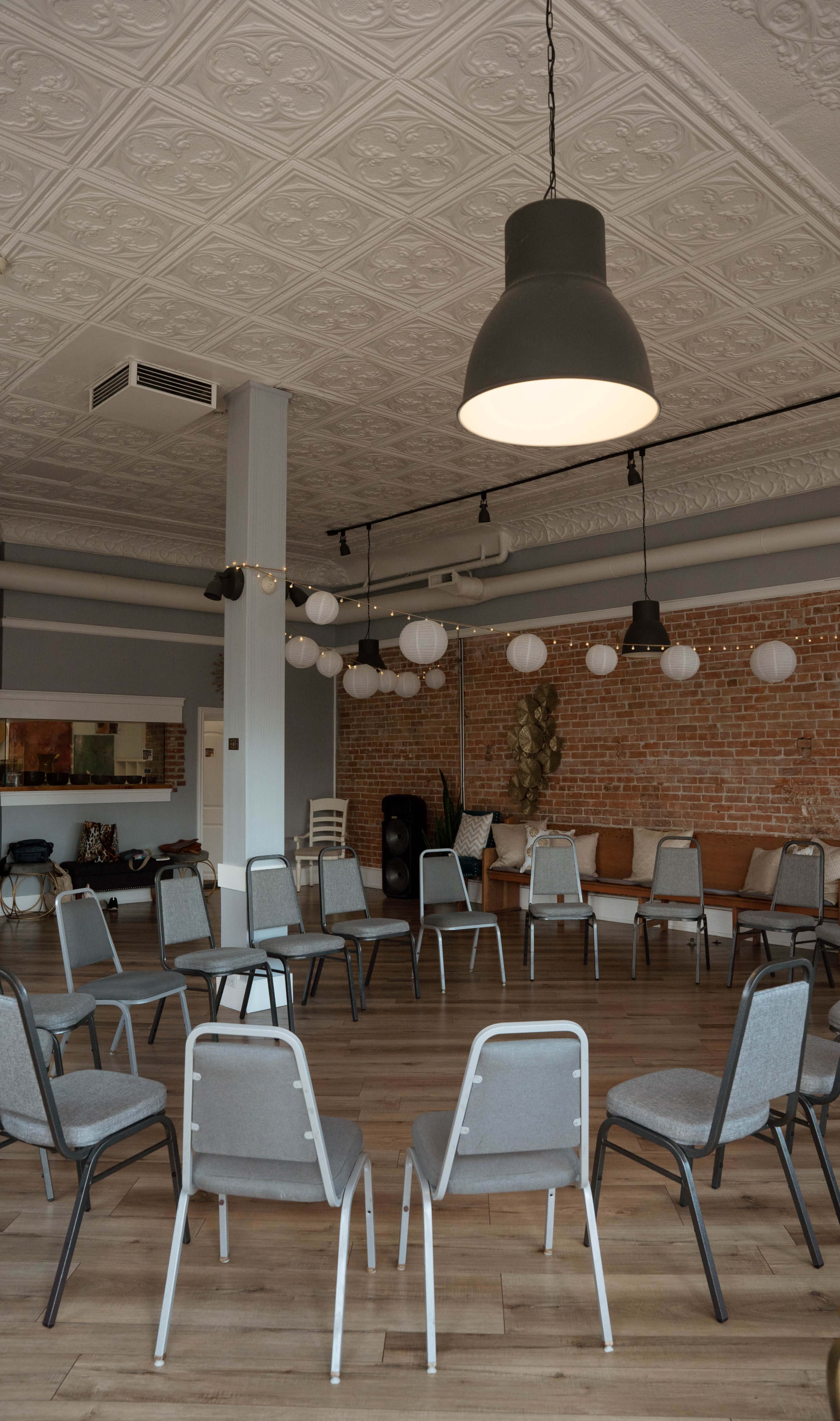 The image shows a circular arrangement of grey chairs in a spacious room with wooden flooring and decorative brick walls.
