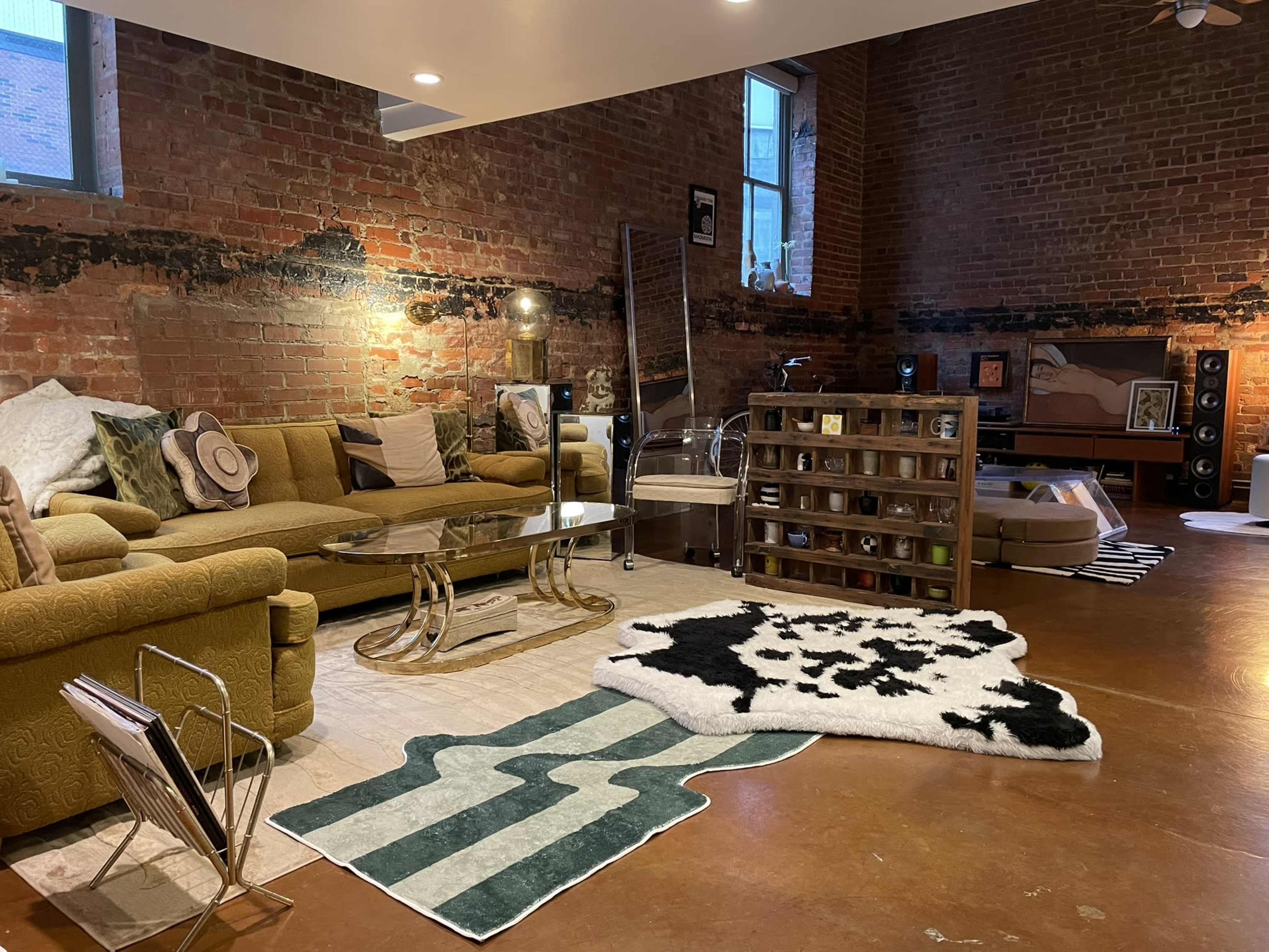 A spacious living room with exposed brick walls, a large green sofa, a glass coffee table, and a cowhide rug, along with shelves displaying various decor items.
