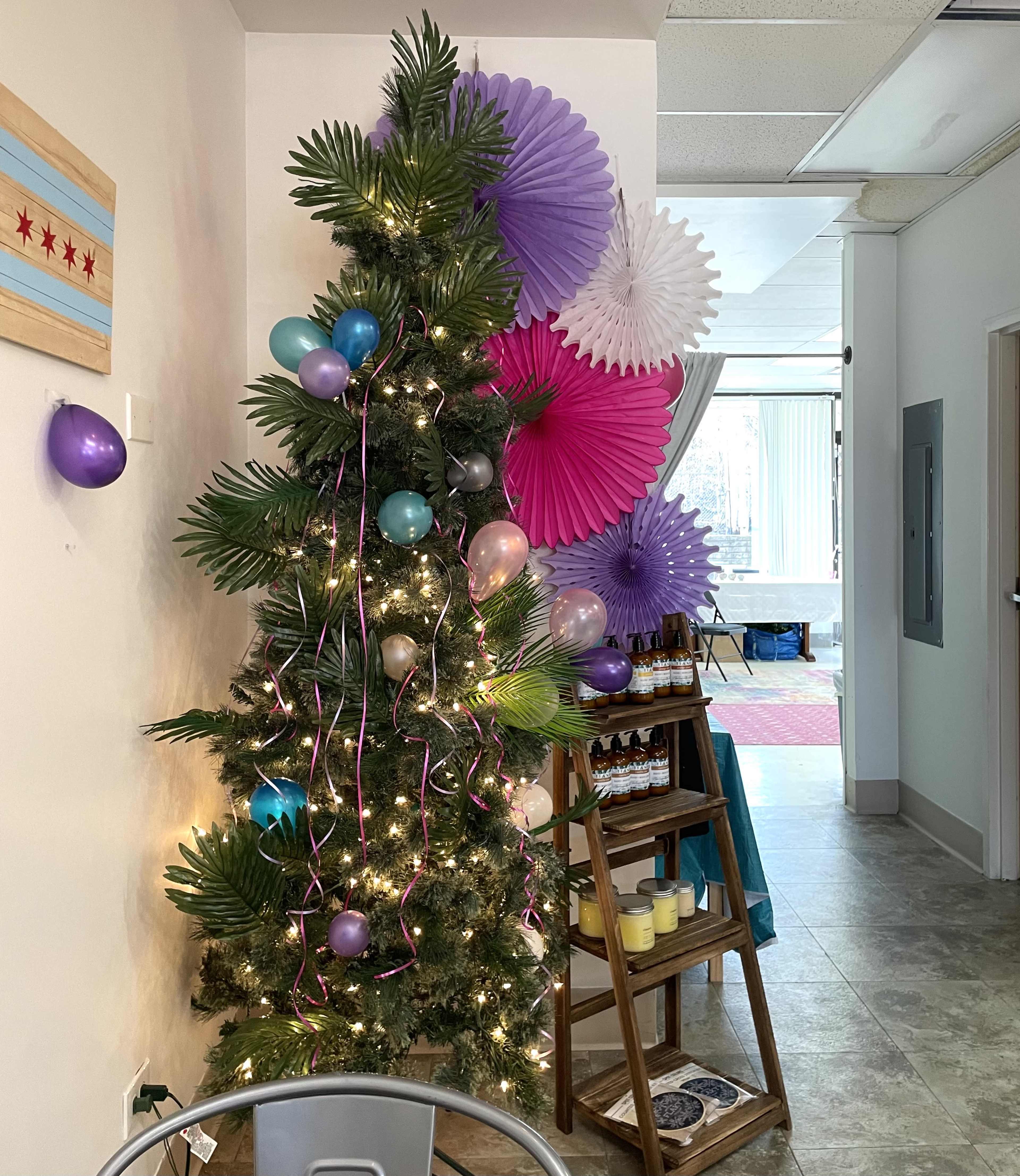 A decorated Christmas tree with colorful balloons and paper fans stands in a well-lit room next to a table displaying jars and candles.