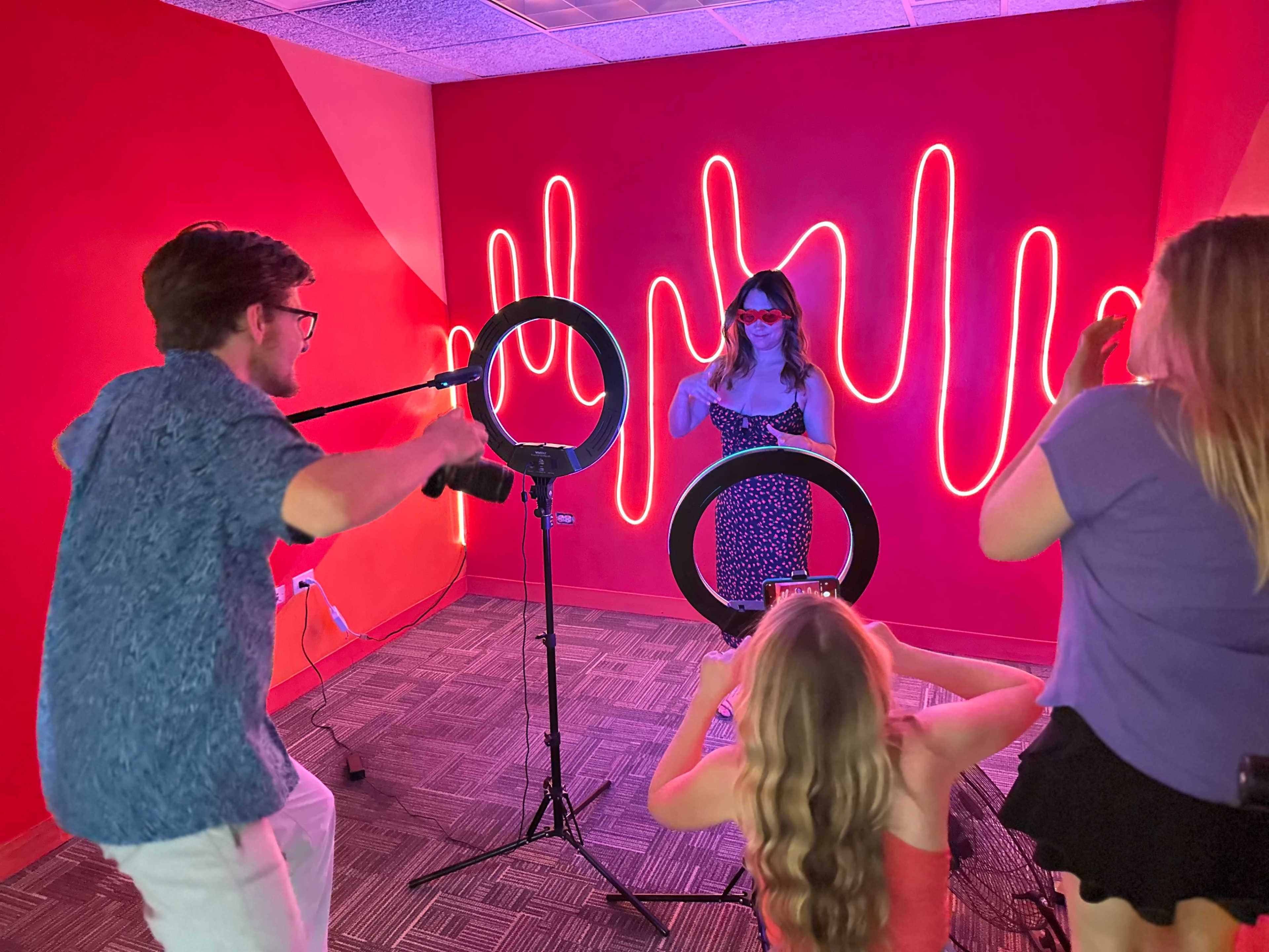 A group of four people is taking photos of a woman posing in front of neon light art in a brightly colored room.