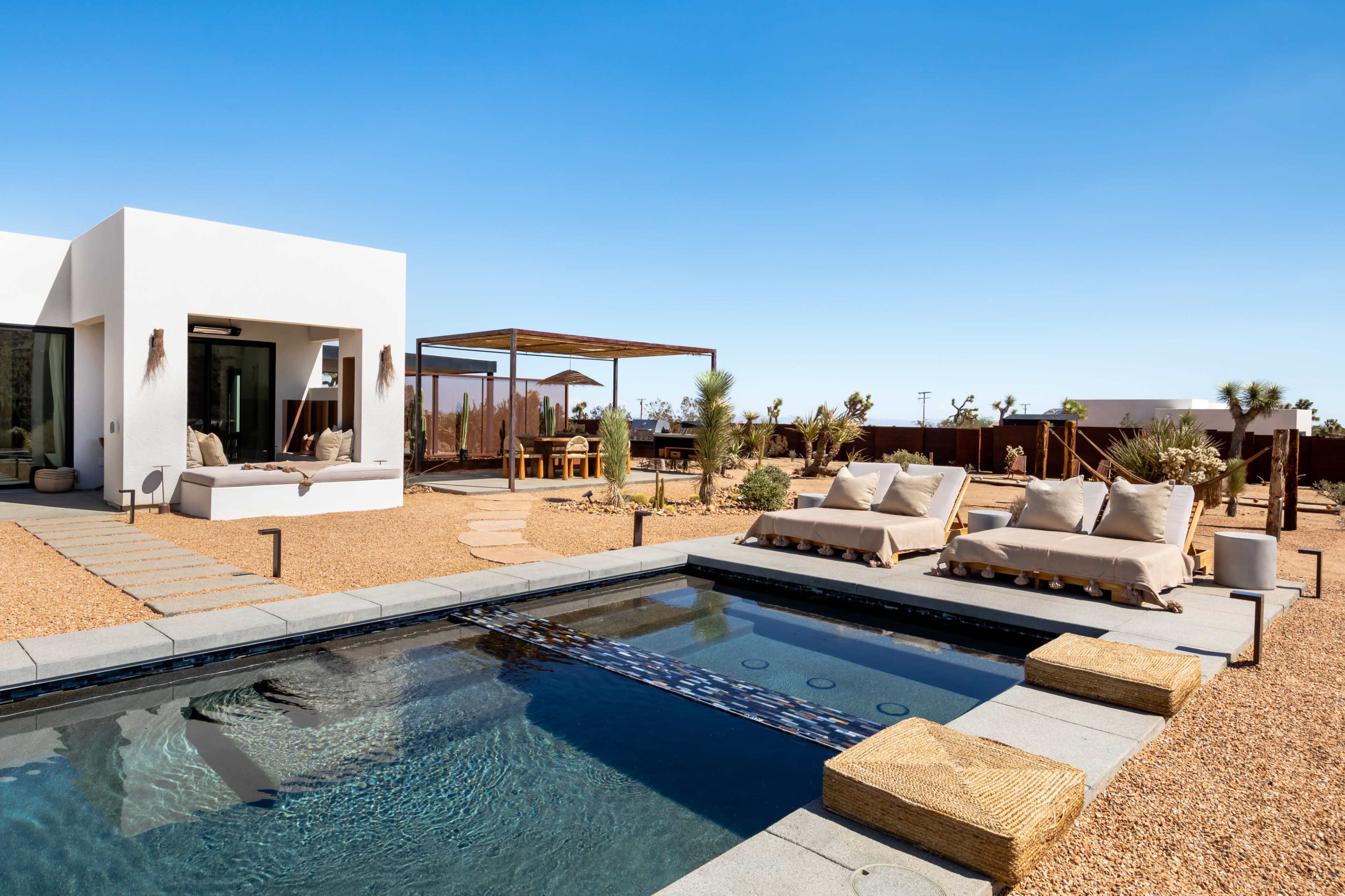 A modern residence features a swimming pool surrounded by lounge chairs and a shaded dining area in a desert landscape.