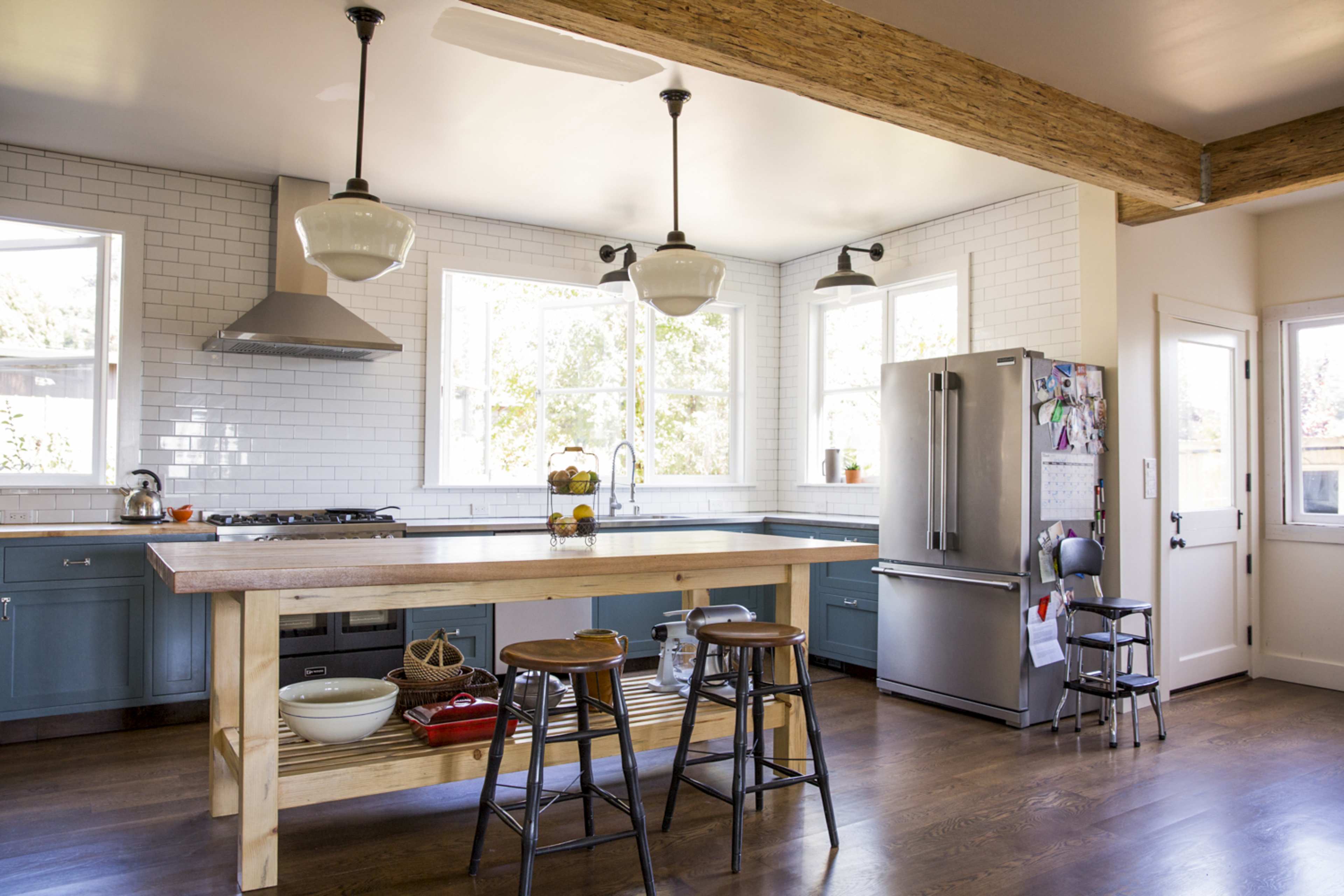 A modern kitchen with blue cabinetry, a large wooden island with stools, and multiple windows allowing natural light to enter.