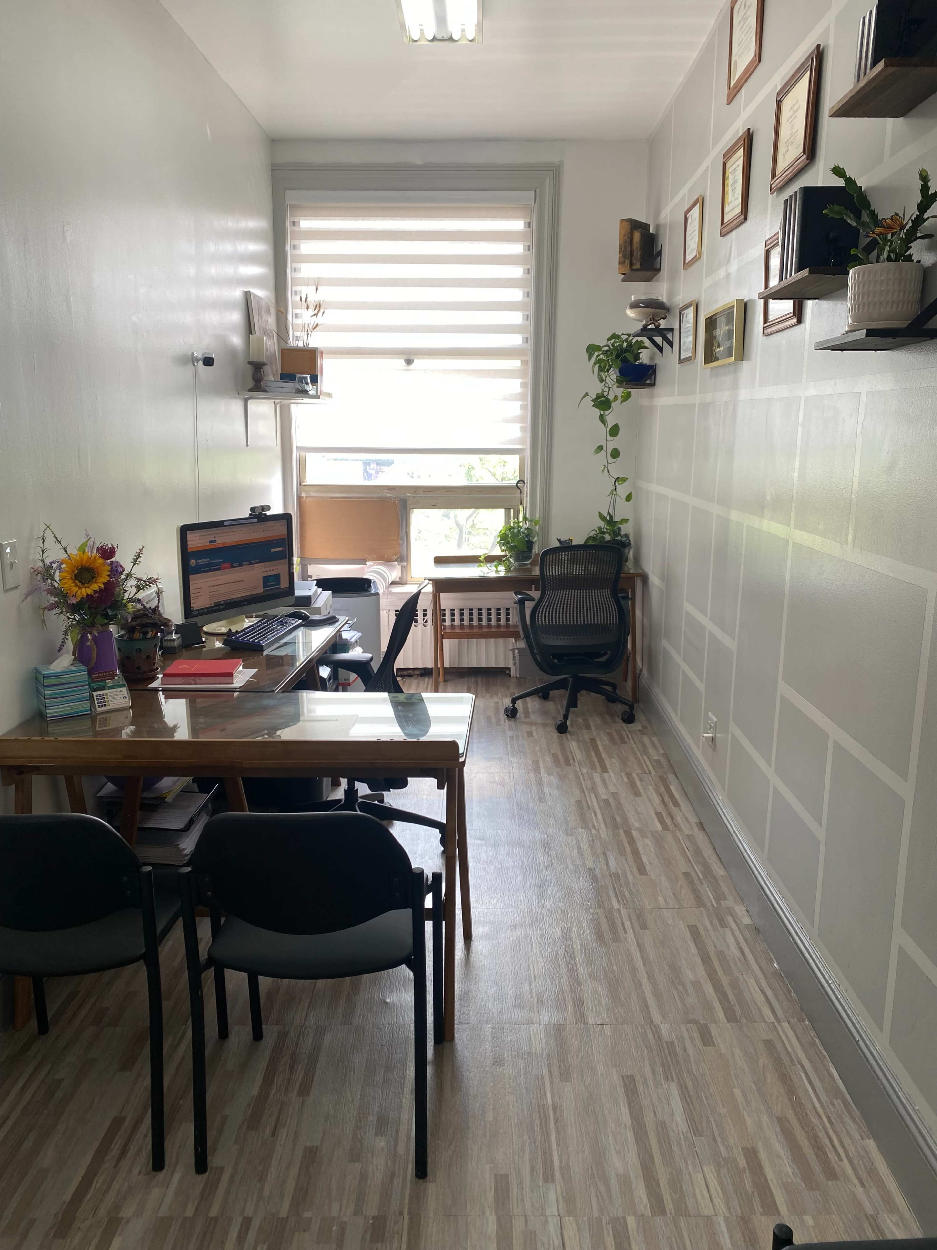 A narrow office space featuring a desk with a computer, a chair, and plants, with walls adorned with shelves and framed documents.