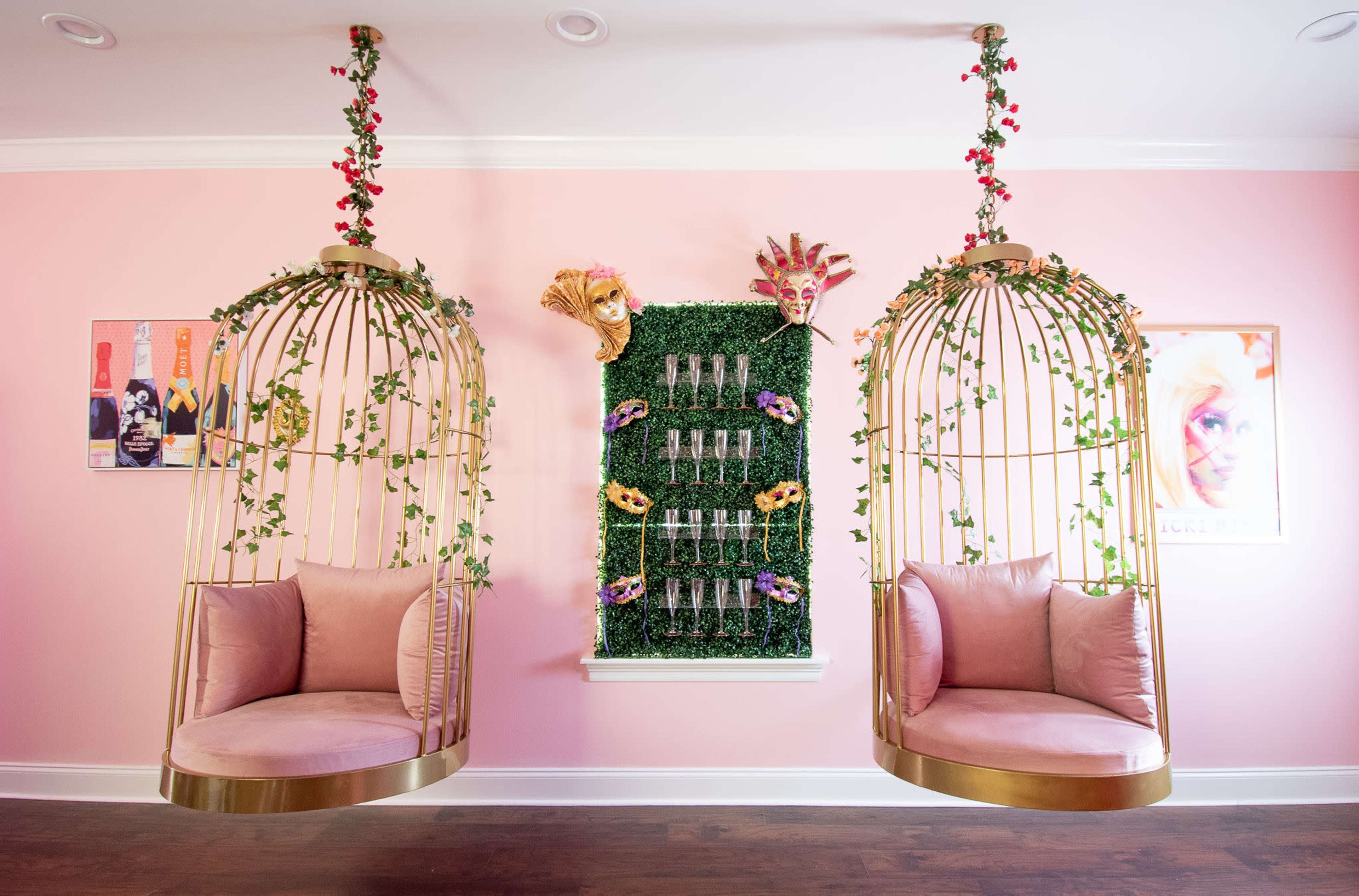 The image features two hanging birdcage chairs adorned with flowers and greenery, flanking a wall decorated with a vertical display of glass champagne flutes and colorful masks against a pink backdrop.