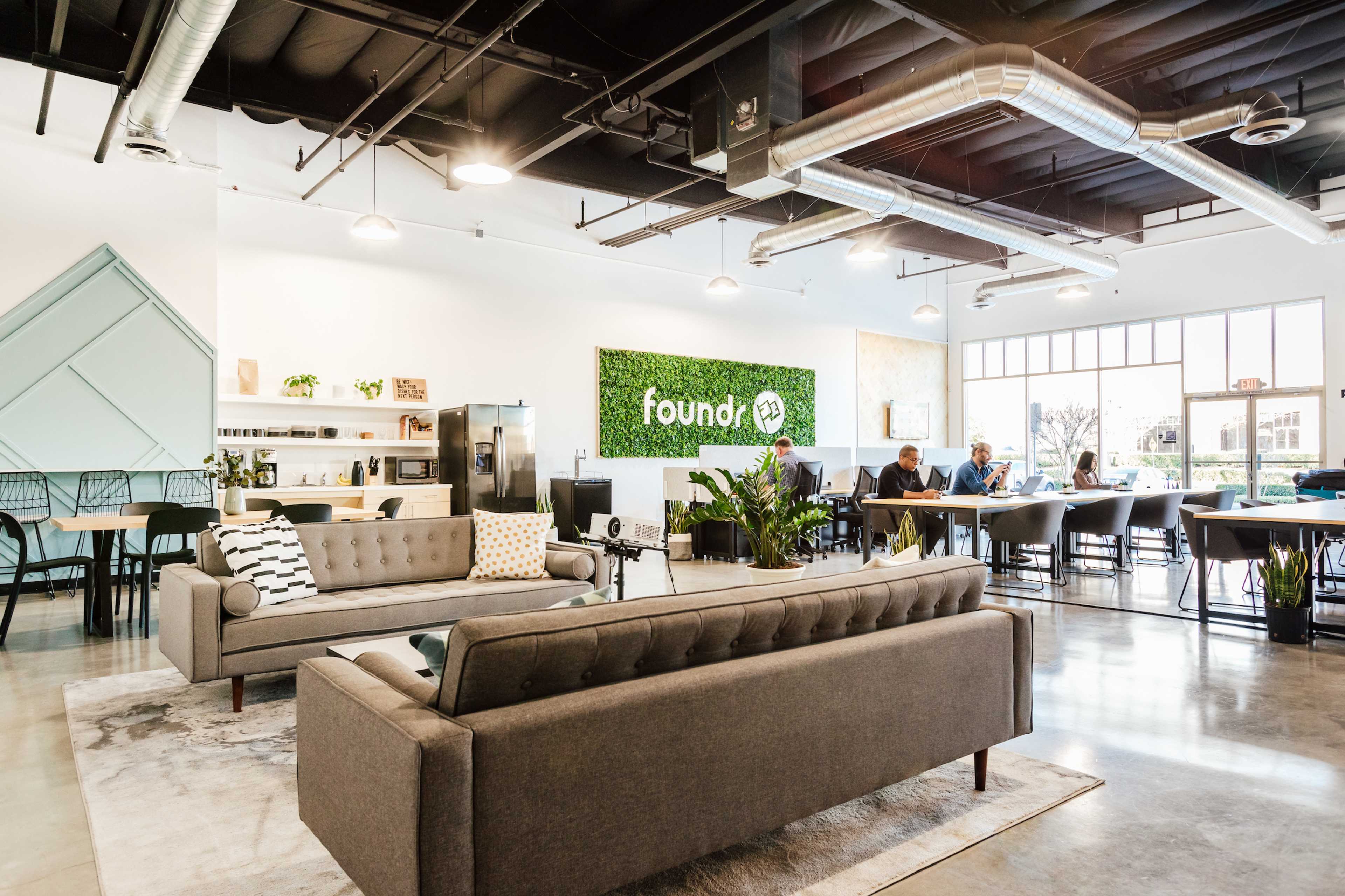 A modern lounge area with gray sofas, a green wall featuring the word "foundry," and several people working at tables in a bright, open space.