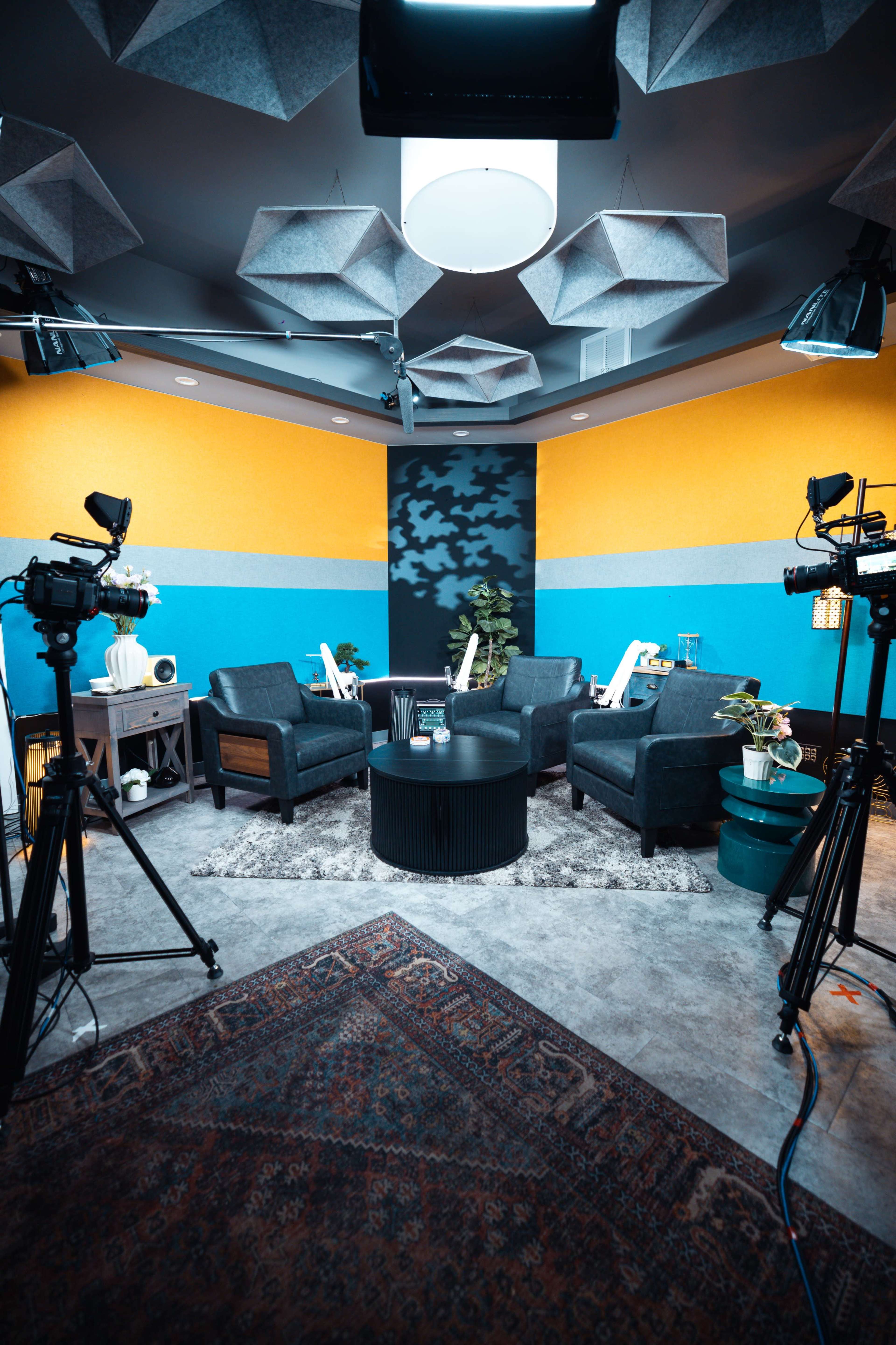 The image shows a modern, well-lit interview studio with gray couches, a round table, and multiple cameras positioned around the space.