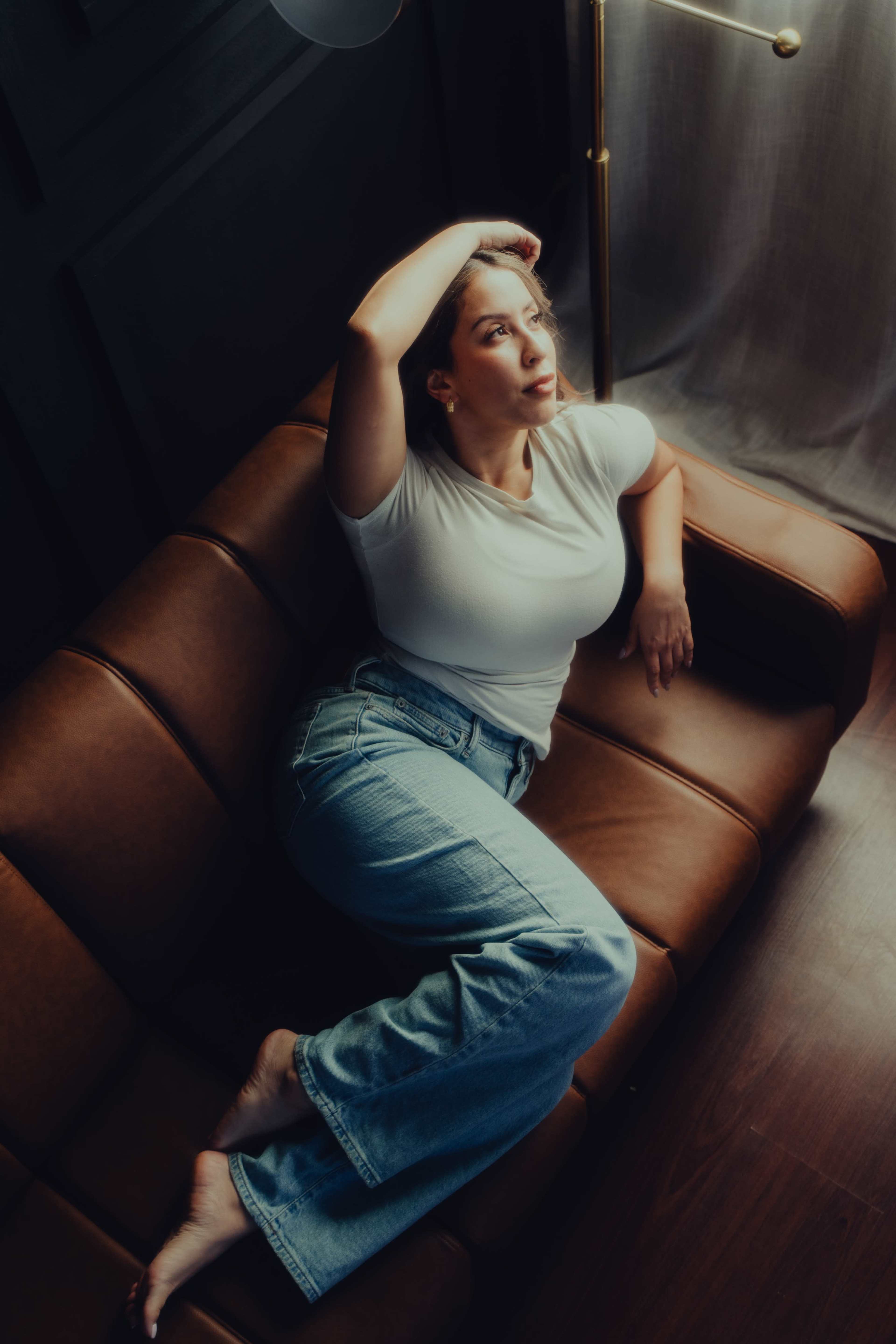 A woman with shoulder-length hair is lounging on a brown leather sofa, gazing thoughtfully out of a window while resting one arm on her head and the other along the back of the couch.
