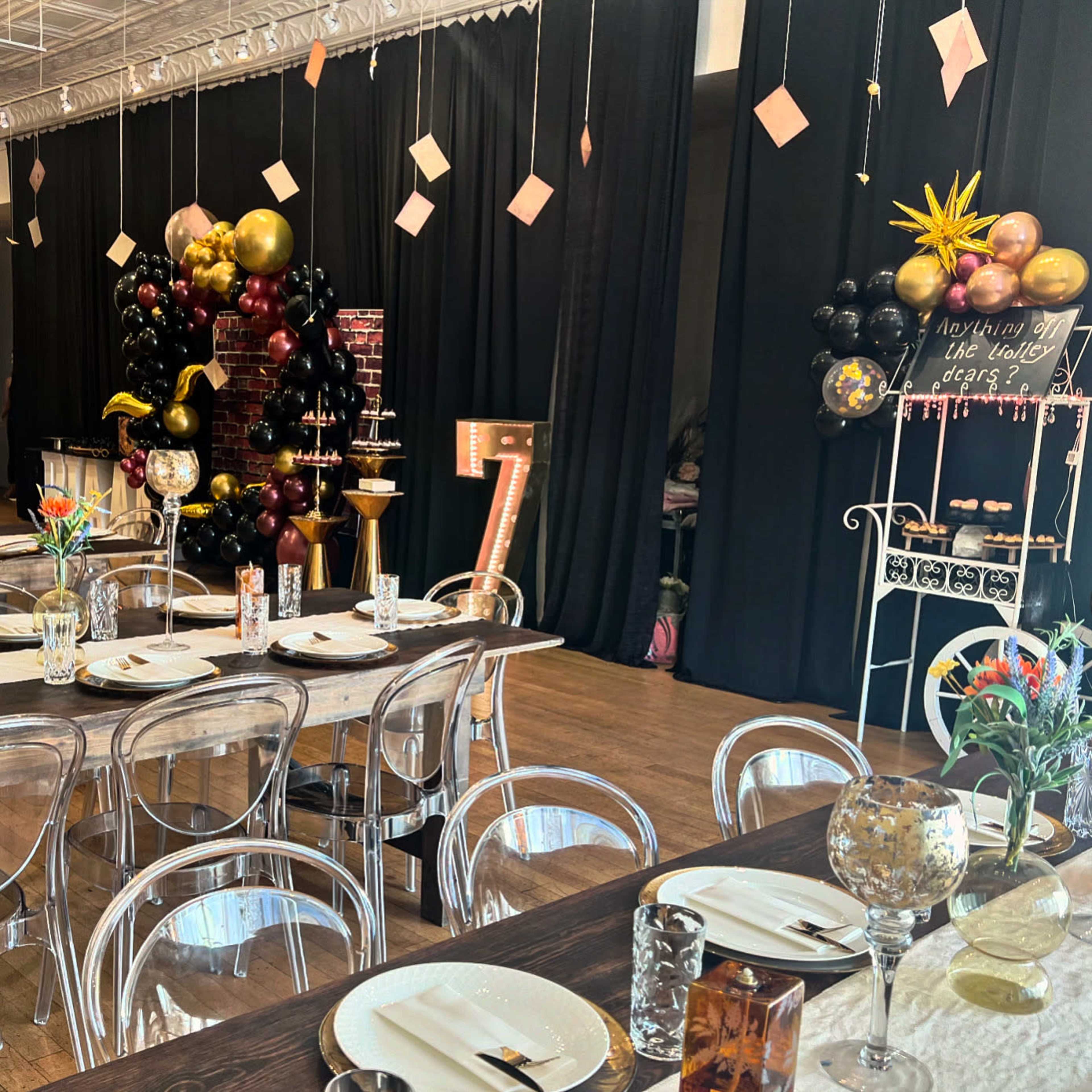 The image shows an elegantly decorated event space with a long wooden table set for dining, surrounded by transparent chairs, and featuring black curtains, colorful balloon arrangements, and decorative elements.
