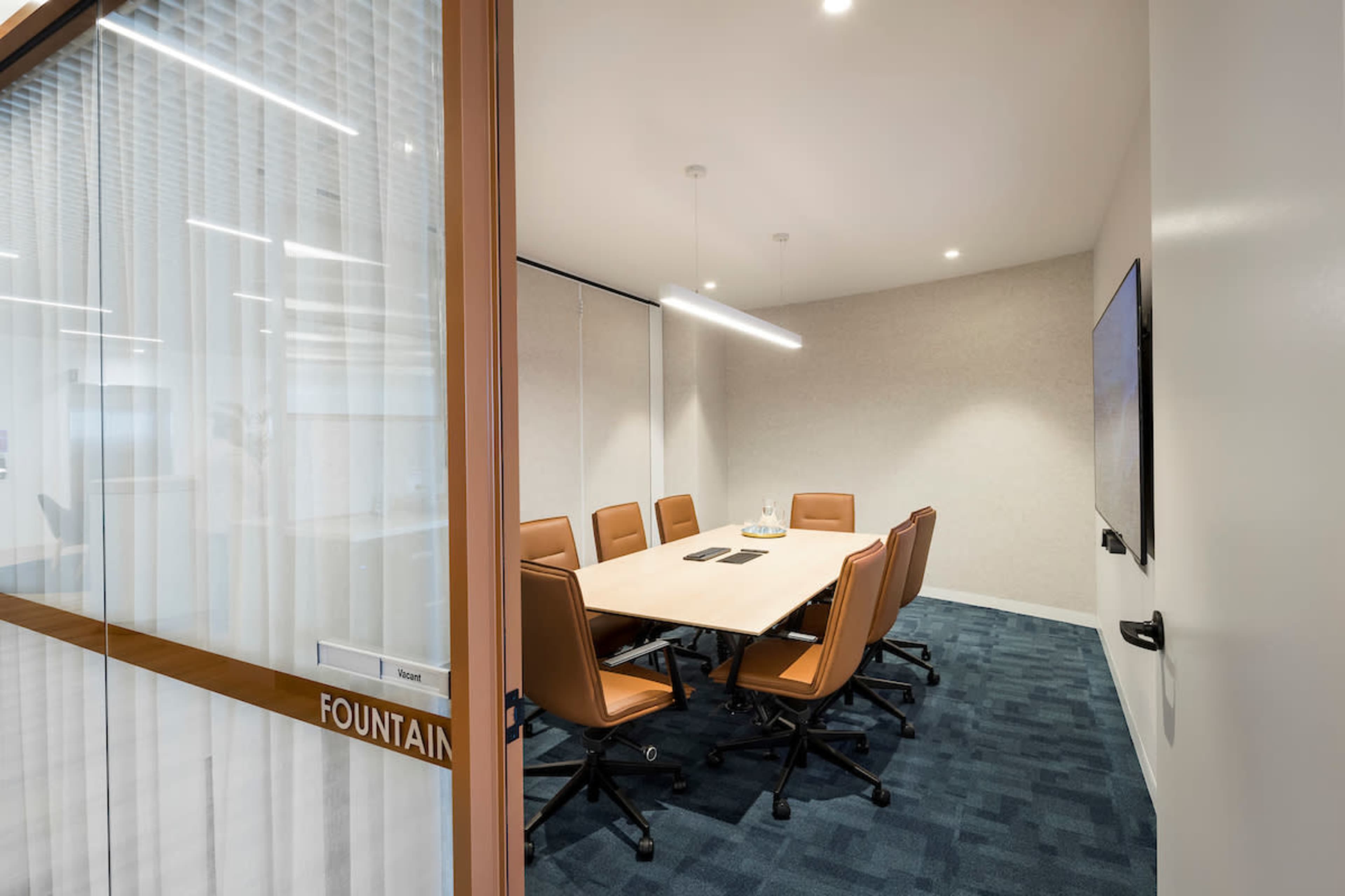 A conference room features a large table surrounded by brown chairs, with a TV mounted on the wall and glass doors labeled "FOUNTAIN."