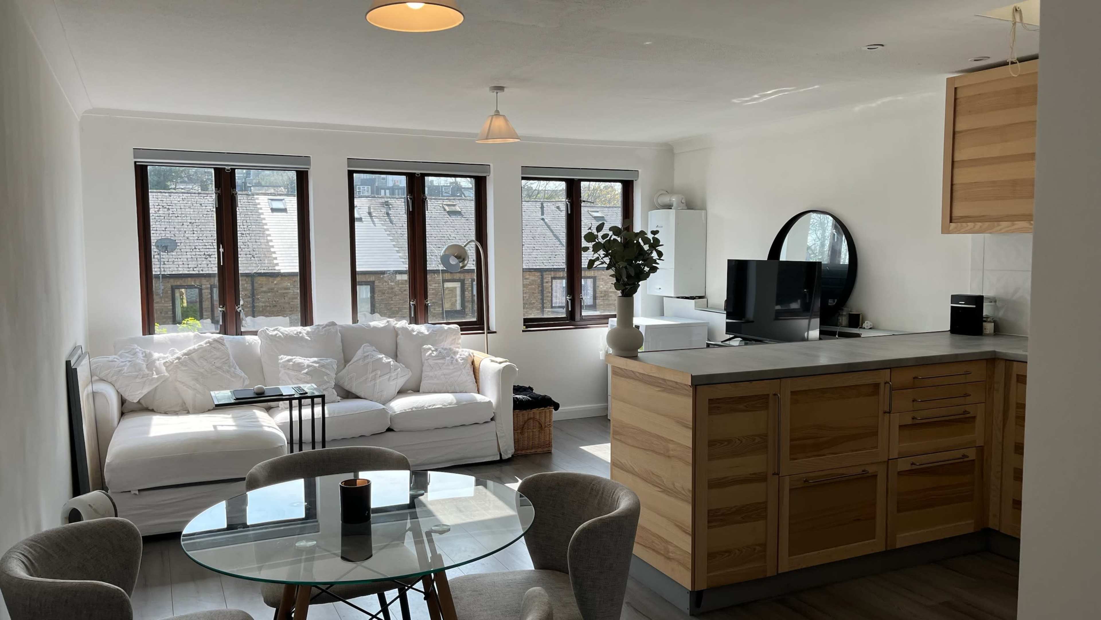 The image shows a modern, open-concept living space with a white couch, a round glass dining table, and a well-equipped kitchen area featuring wooden cabinets.