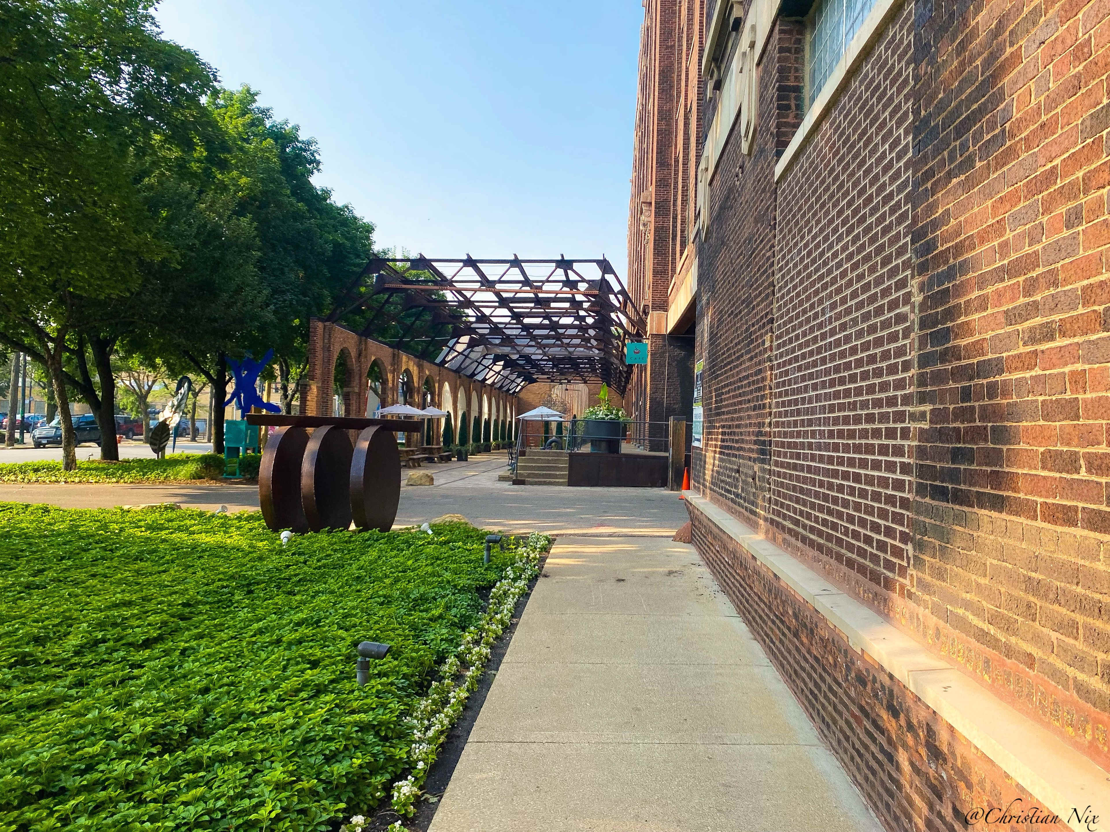 The image shows a brick building beside a pathway lined with greenery and a decorative art installation in the foreground.