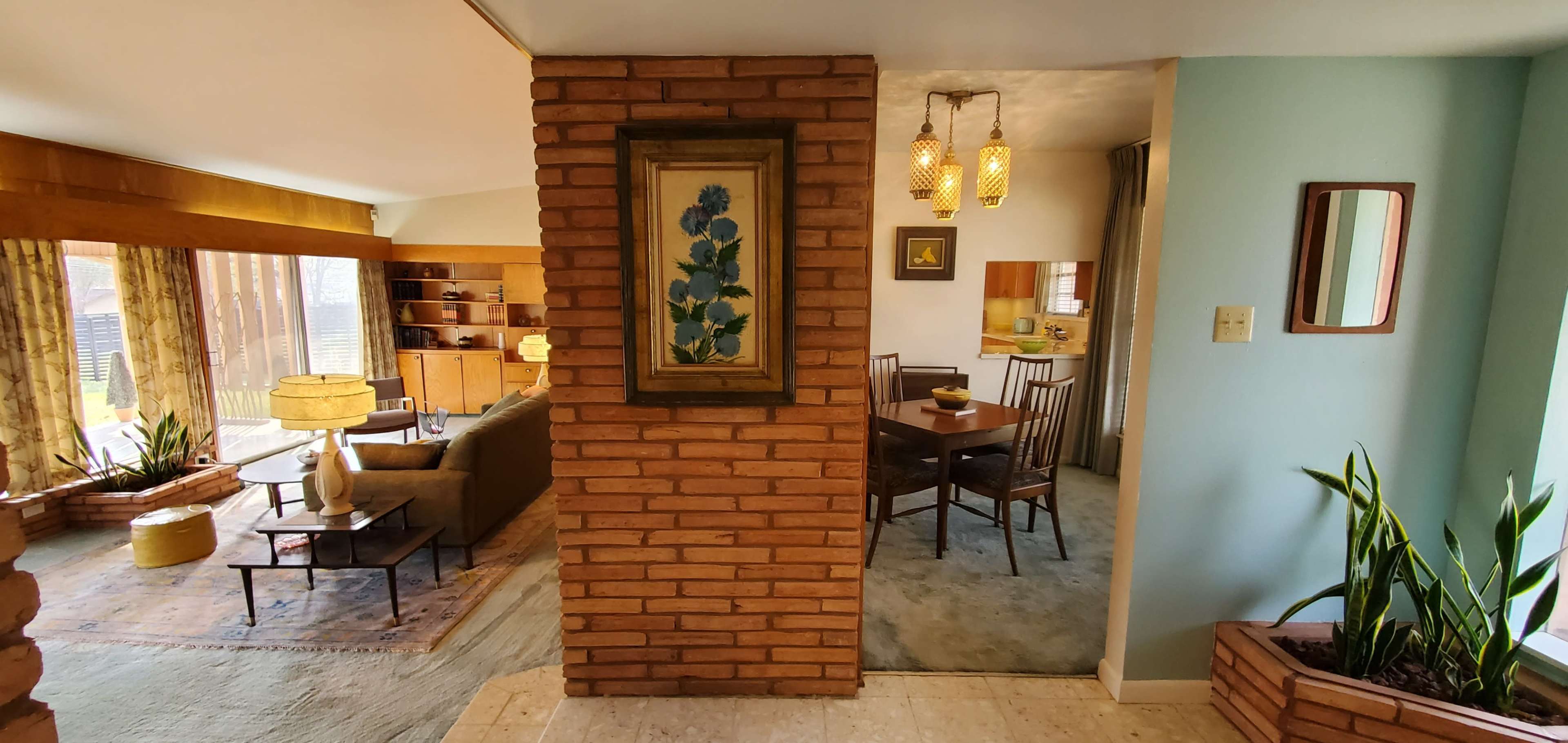 A split-level interior featuring a brick wall with a framed painting, leading to a dining area visible through an open layout, with a living room furnished in the background.