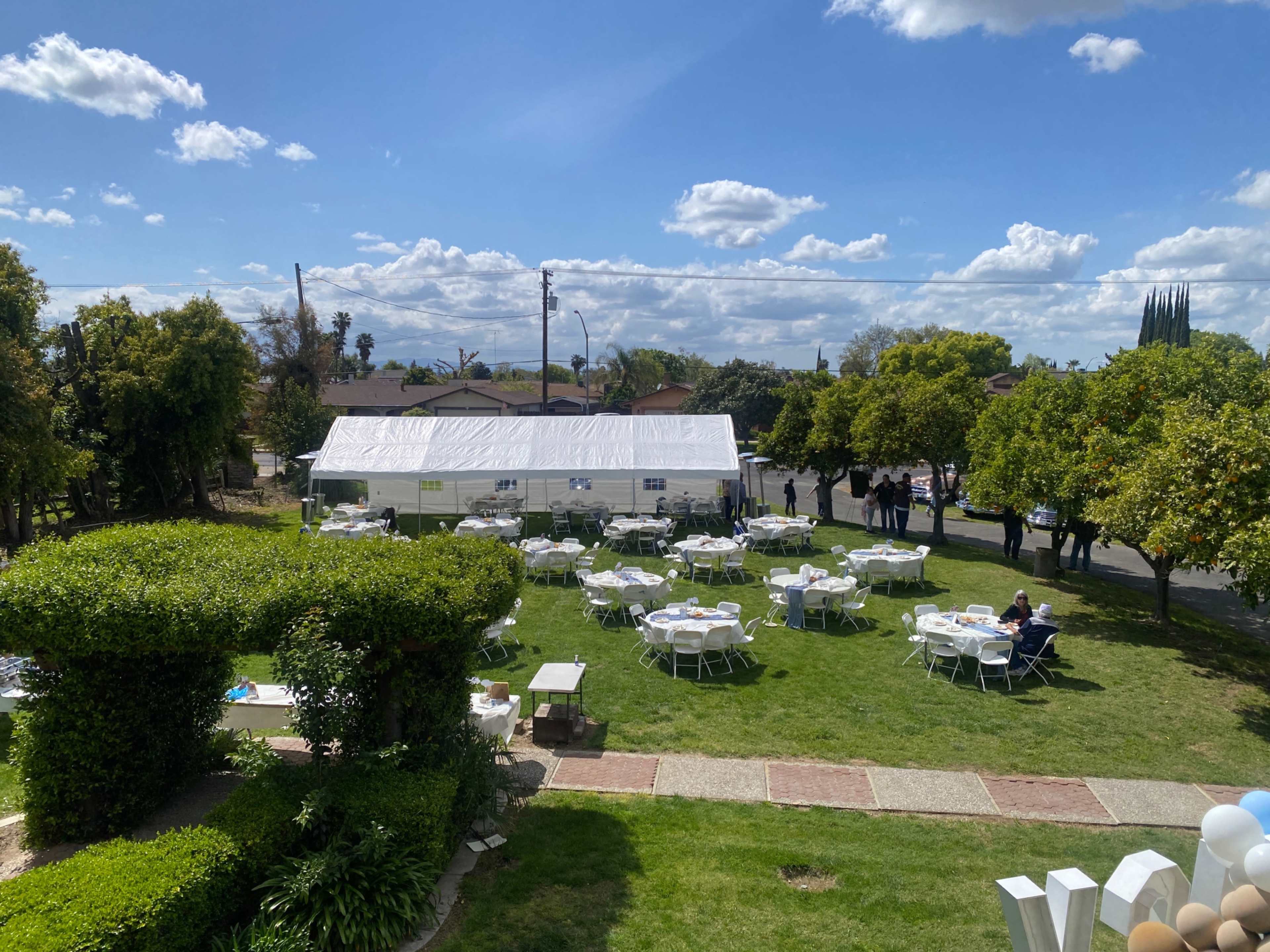 A large white tent is set up in a grassy area surrounded by trees, with multiple tables and chairs arranged for an outdoor event.