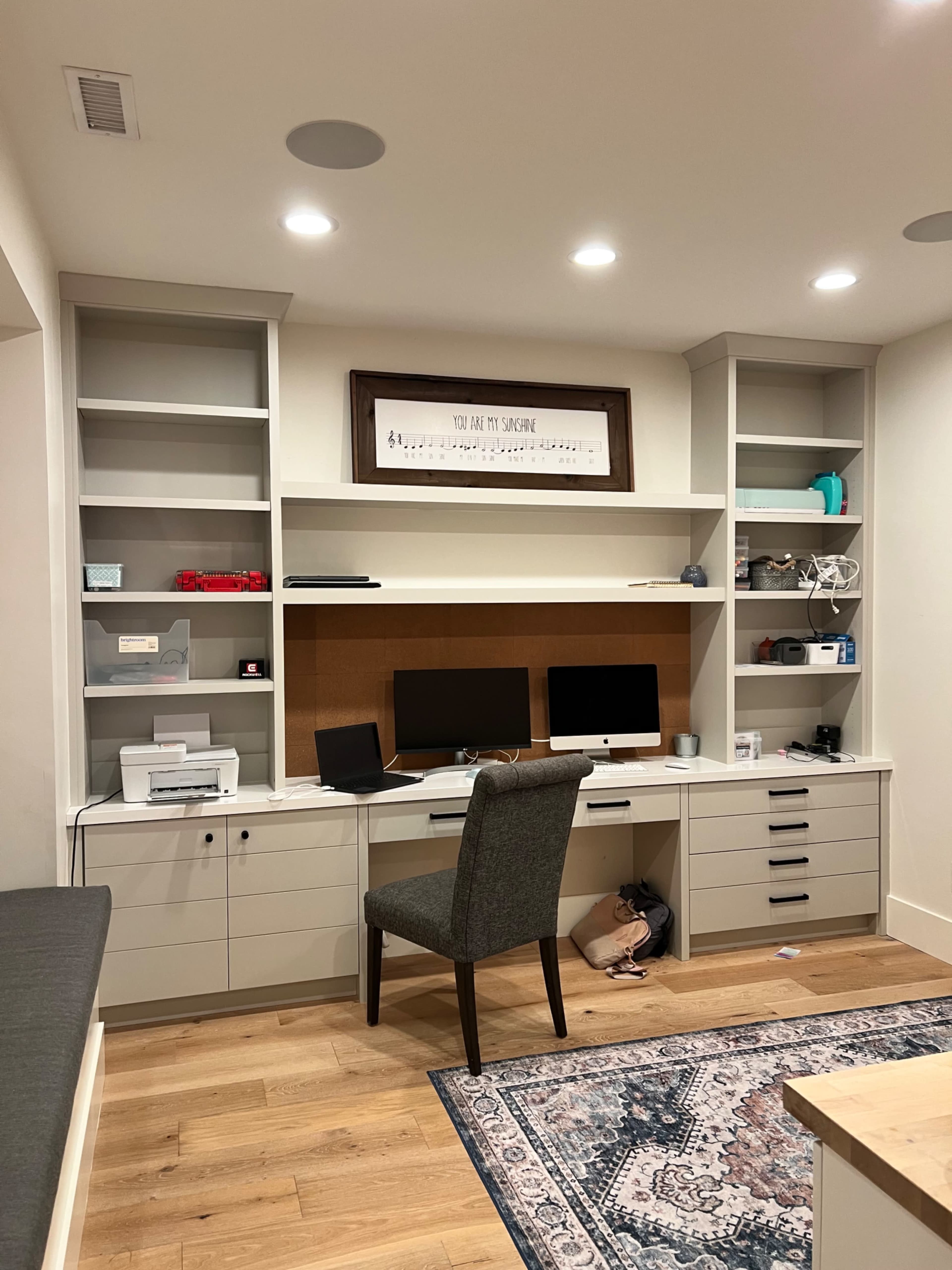 The image shows a modern home office with a built-in desk, two computer monitors, a chair, and various organizational shelves.