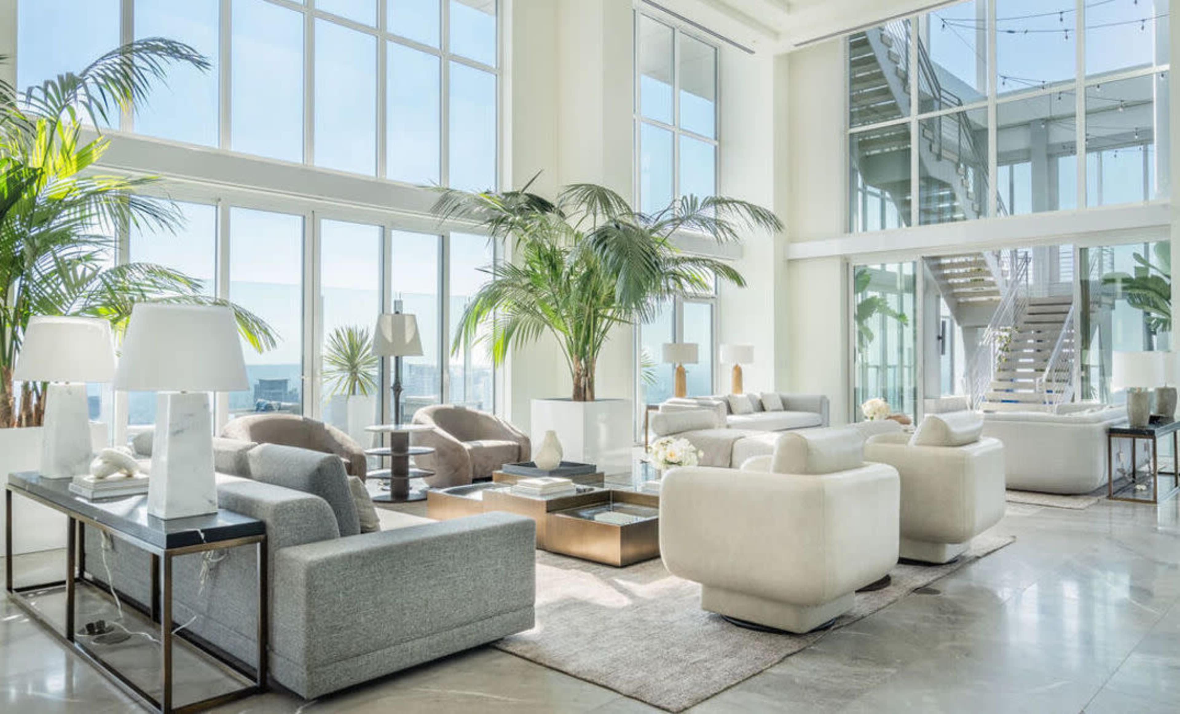 A spacious modern living area features large windows, tropical plants, and stylish furniture arranged around a central coffee table.