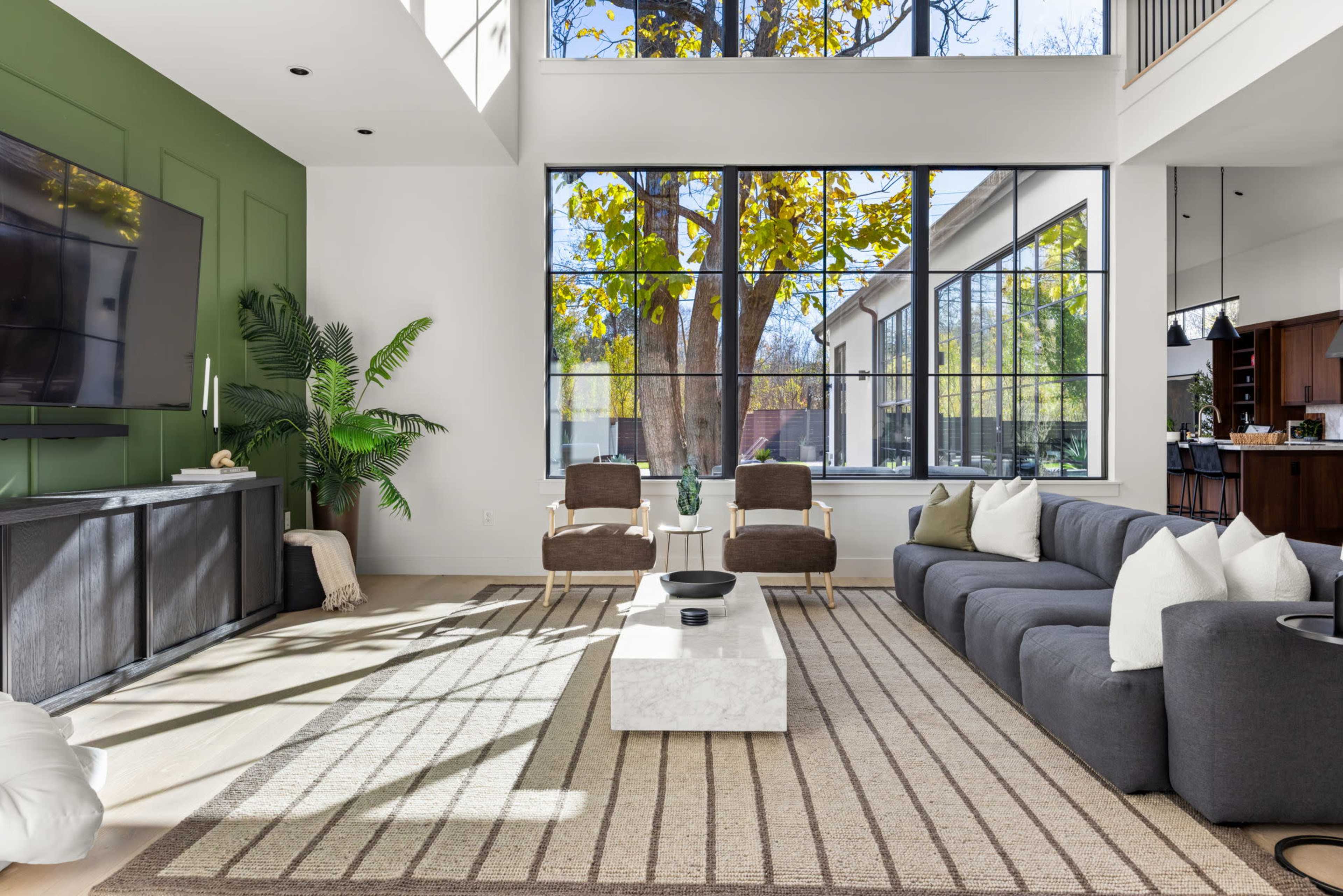 The image shows a modern living room with a large window overlooking trees, featuring a gray sectional sofa, two brown chairs, a coffee table, and a television mounted on the wall.